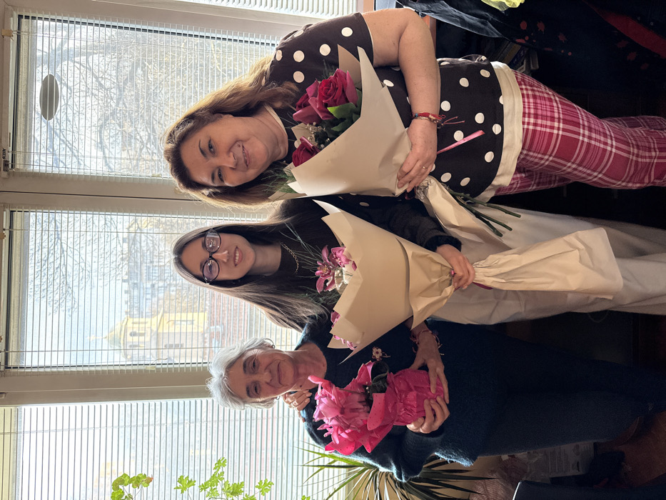 Three females of varying ages are standing vertically indoors, positioned in front of a window fitted with horizontal blinds. The subject on the left is an elderly woman with grey hair, wearing a dark long-sleeved top, holding a potted pink cyclamen wrapped in bright pink paper. Her left arm is positioned behind the middle subject. The central subject is a younger woman with long dark hair and eyeglasses, wearing a dark long-sleeved top and light-colored pants. She holds a bouquet of light pink and purple flowers wrapped in plain brown paper. The subject on the right is a middle-aged woman with shoulder-length brown hair, wearing a dark brown short-sleeved top with white polka dots and pink plaid pants. She holds a bouquet of red roses wrapped in plain brown paper and displays a multi-strand bracelet on her right wrist. All three individuals are smiling towards the camera. Through the window, the golden dome and clock tower of the Central Mineral Bathhouse, now the Regional History Museum - Sofia, are clearly visible, identifying the location as Sofia, Bulgaria, likely from an upper floor of an adjacent building. A green potted plant is partially visible on the far left of the frame.
