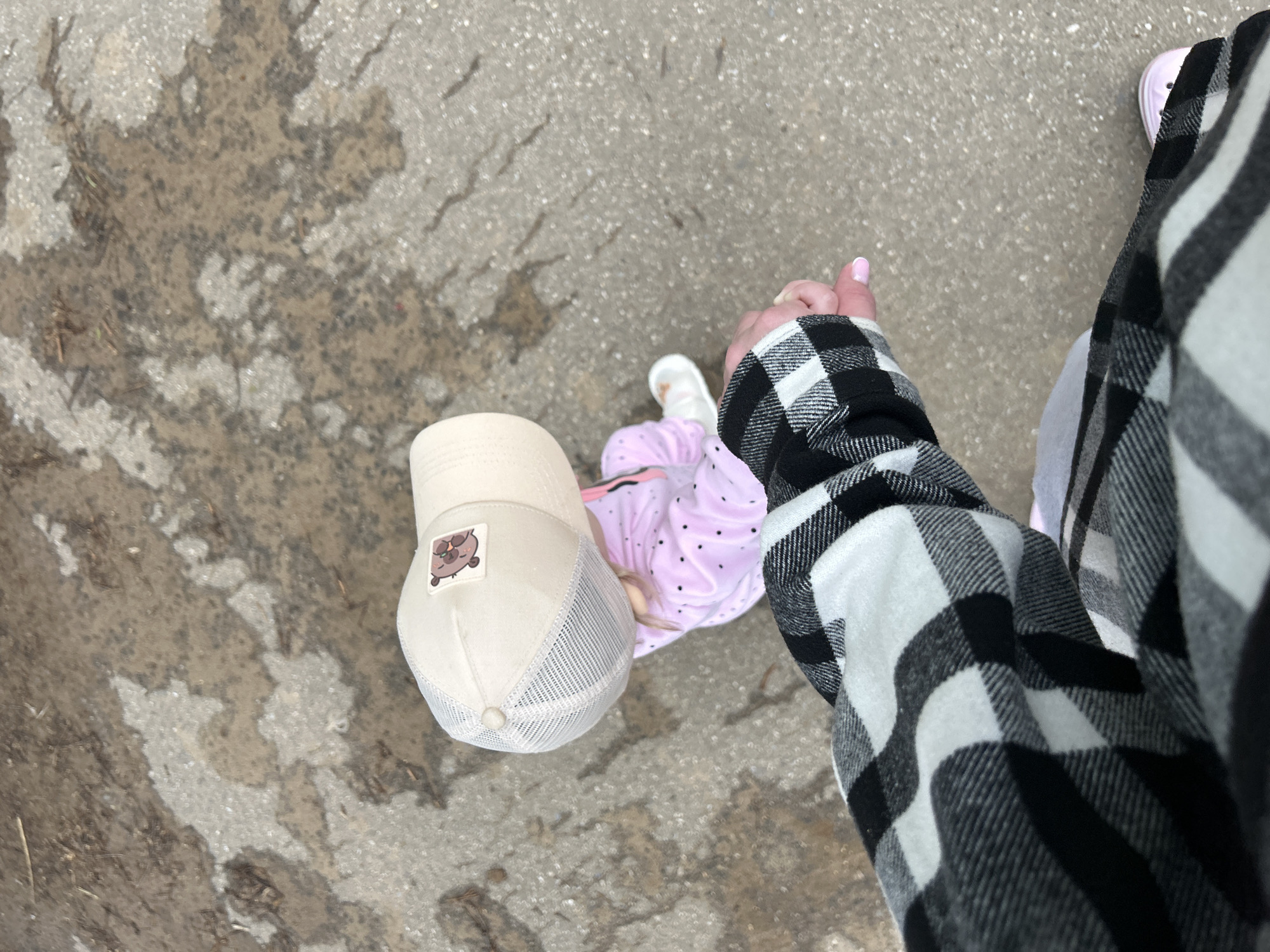 An adult's hand, with painted pink fingernails, holds the hand of a young child. The adult wears a black and white plaid shirt. The child is wearing a light pink outfit with dark polka dots and a light-colored baseball cap with a bear patch. They are standing on a wet, gray, asphalt-like surface with some debris. The scene appears to be outdoors. The location in Chaikovsky, Russia cannot be determined from the image.