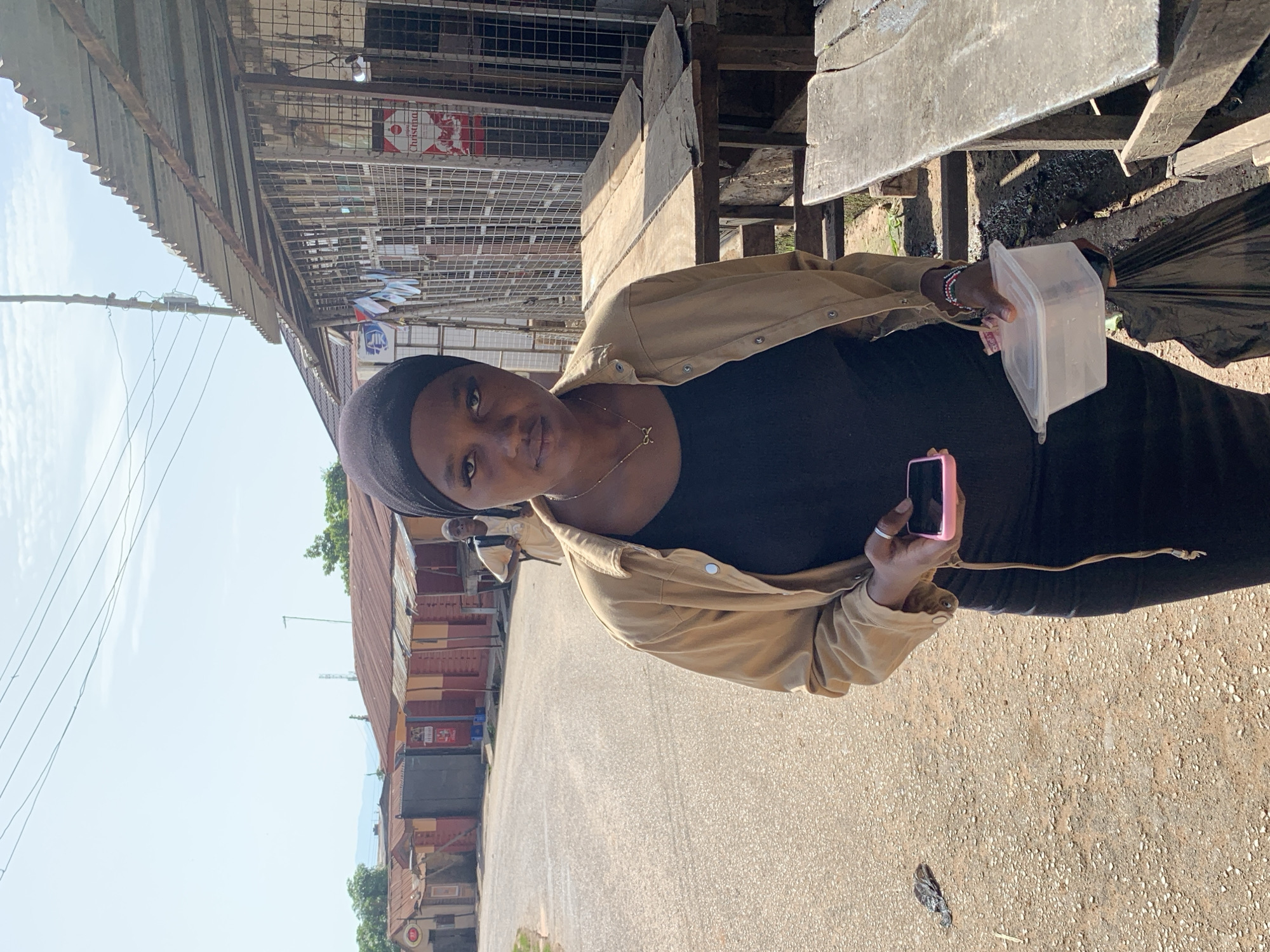 A young Black woman wearing a black hijab and a light brown jacket over a black dress is standing on a dirt road in Jos, Nigeria. She is holding a pink smartphone in her right hand and a clear plastic container in her left. Behind her, a younger Black person wearing a light-colored t-shirt and backpack walks away from the camera. The background features several low-rise buildings with corrugated metal roofs, some appearing to be market stalls. The sky is bright and overcast.