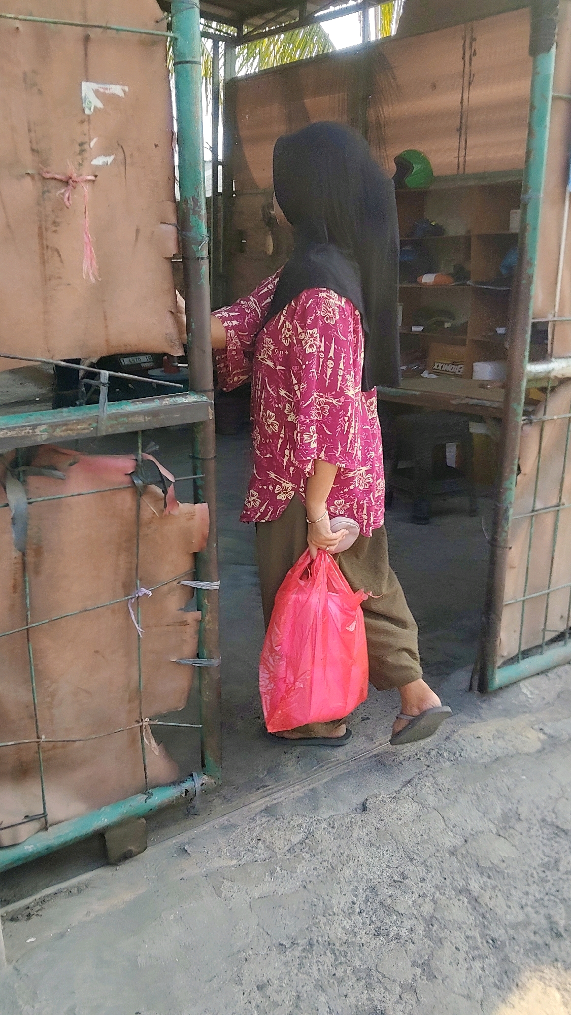 In Mekarmukti, Indonesia, a person wearing a black hijab and a pink patterned shirt is walking through a doorway. They are carrying a bright pink plastic bag in their right hand and appear to be wearing sandals. The environment includes a metal gate with green painted posts, a rough brown material on the left, and shelving units in the background. The ground is made of concrete with visible cracks and texture. The lighting suggests it is daytime, with sunlight illuminating parts of the scene.