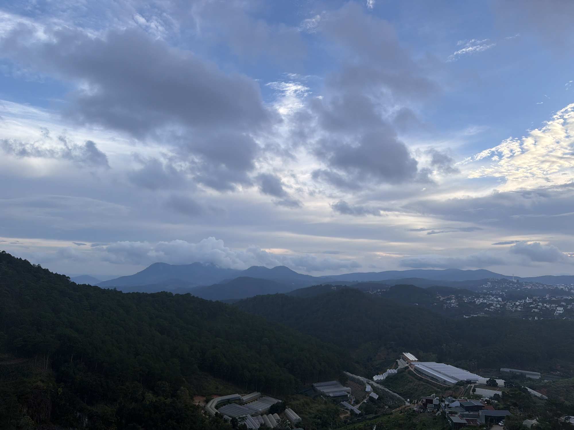 The image displays a panoramic view of a mountainous landscape, likely in or around the city of Xuân Thành, Vietnam. In the foreground, a dense forest covers a steep hillside, dotted with several greenhouses and some scattered residential buildings. A winding road is visible on the left side of the hill.
Further into the distance, a series of rolling hills recedes, covered in a similar dark green vegetation. The farthest hills are veiled in a hazy, blue-grey tone, suggesting a considerable distance and possibly a lower elevation or atmospheric conditions.
The sky above is dramatic, filled with a mix of dark, heavy clouds and patches of brighter, illuminated sky, possibly indicating the approach or departure of a storm, or the golden hour around sunrise or sunset. The light filtering through the clouds creates a dynamic and moody atmosphere. A small, distant town or city is visible on the right-hand side, nestled in the valleys between the hills, characterized by numerous white rooftops.
No people are discernible in the image. The scene appears to be a natural landscape with human settlements and agricultural structures integrated into it. The overall impression is one of a vast, undulating terrain under a dynamic sky.