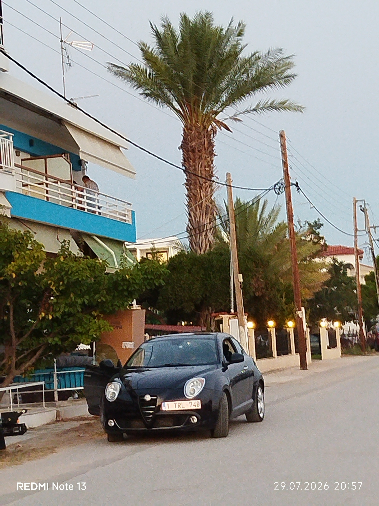 This image captures a street scene in Sithonia Municipal Unit, Greece, at dusk. A black Alfa Romeo MiTo car, with its driver's side door open, is parked on the right side of the road. The car's license plate reads "1 TRL 748." In the background, a multi-story building with a blue facade and balconies is visible, with a palm tree standing tall beside it. Further down the street, another building with a red-tiled roof is present, and a row of illuminated posts lines the sidewalk. The sky is overcast, suggesting twilight. The timestamp "29.07.2026 20:57" is displayed in the lower right corner, along with "REDMI Note 13" in the lower left. A person is visible on a balcony of the building on the left.