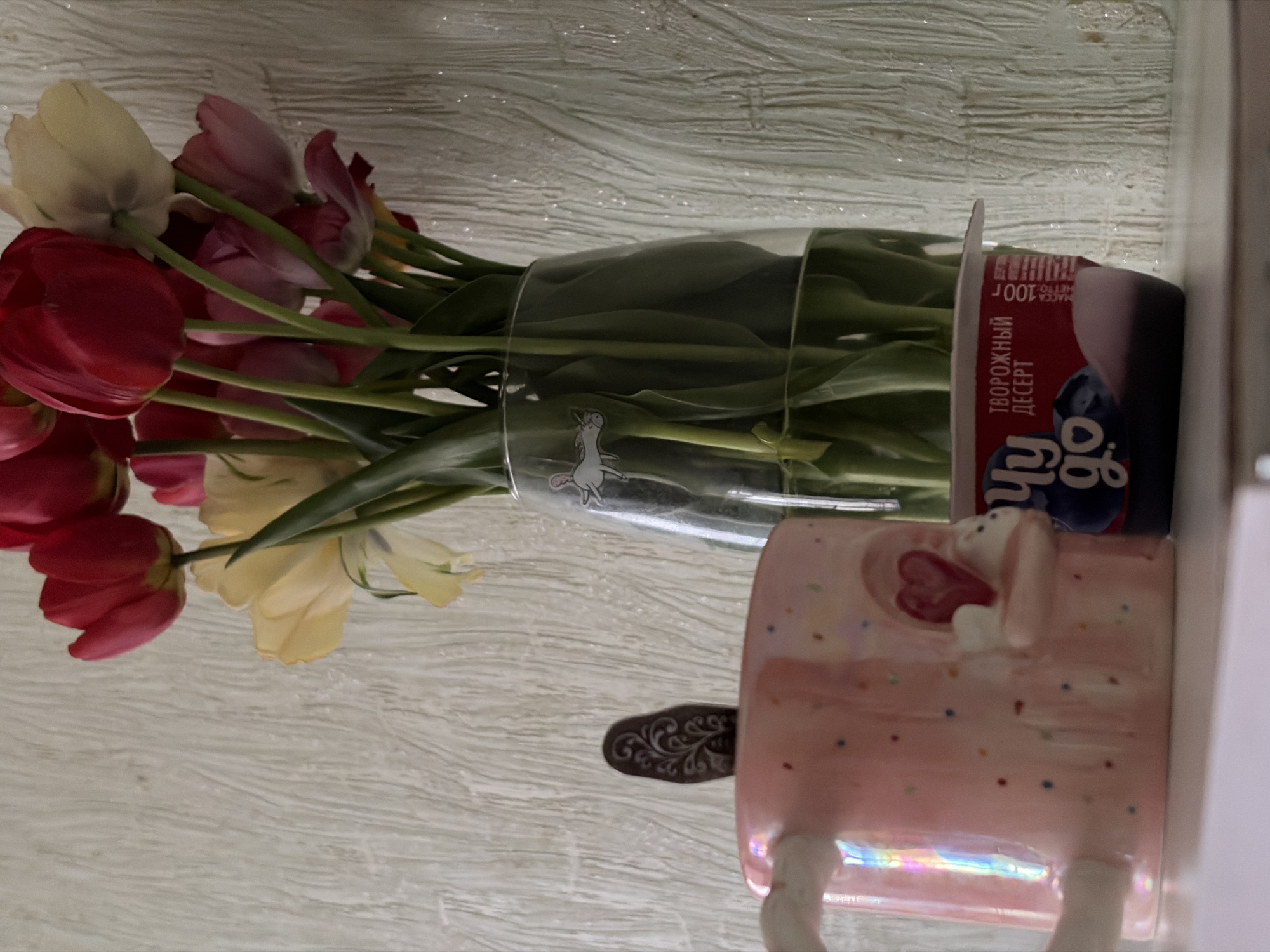 The image shows a bouquet of tulips in a glass jar, placed next to a pink mug decorated with a bunny and heart design. The jar appears to be repurposed from a yogurt or dessert container, with Russian text visible on the label. There's also a decorative spoon visible near the mug. The background is a textured white surface, possibly a wall or tabletop. The location, Chaikovsky, Russia, is not discernible from the image itself.