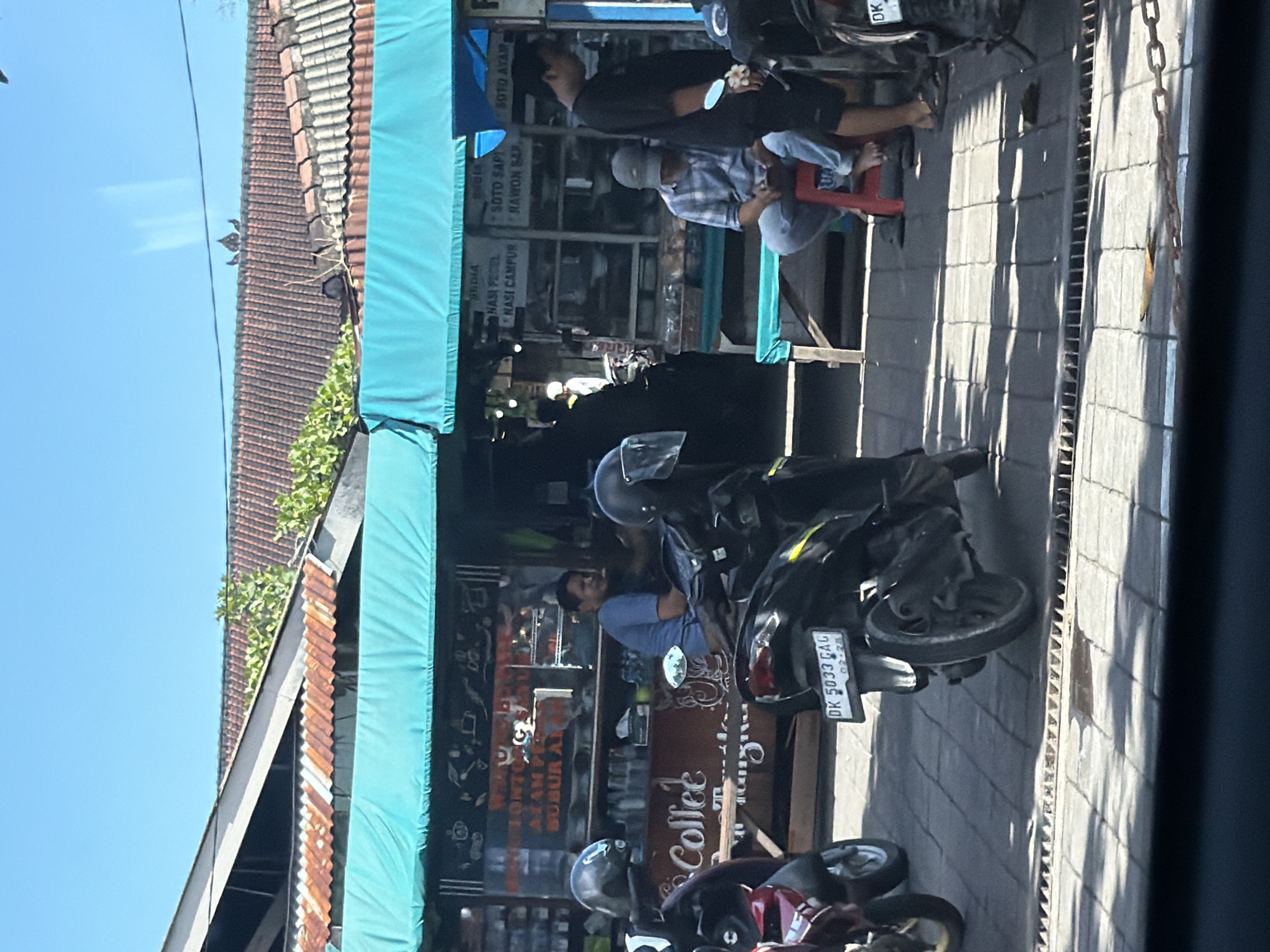This image captures a vibrant street scene in North Kuta, Indonesia, bathed in bright daylight under a clear blue sky. The setting appears to be a casual roadside eatery or warung, shaded by a turquoise blue tarp canopy.
Several men are present, engaged in various activities. One man in a black shirt stands beside a motorbike, while another, wearing a plaid shirt and cap, is seated on a red stool, looking down. Further into the stall, a man in a blue shirt is visible near a counter. These individuals contribute to the bustling atmosphere of a local gathering spot.
A prominent feature of the scene is the array of parked motorbikes, a common mode of transport in Indonesia. One motorbike's license plate, "DK 5033 GAG," is clearly visible. The eatery features a counter and various signs. The street itself is paved, and strong, defined shadows are cast across the ground, indicating direct sunlight and a clear day. In the background, traditional tiled roofs of buildings and some tropical greenery are visible.
Visible text on the signs advertises local Indonesian cuisine, including "SOTO AYAM" (chicken soup), "SOTO SAPI" (beef soup), "RAWON SAPI" (beef black soup), "NASI PECEL" (rice with mixed vegetables and peanut sauce), and "NASI CAMPUR" (mixed rice). A sign for "Coffee" is also prominent. Other partial text includes "BONT," "ALAM," and "BUBUR AR," suggesting more menu items. The scene encapsulates a typical, active street food environment in Indonesia.