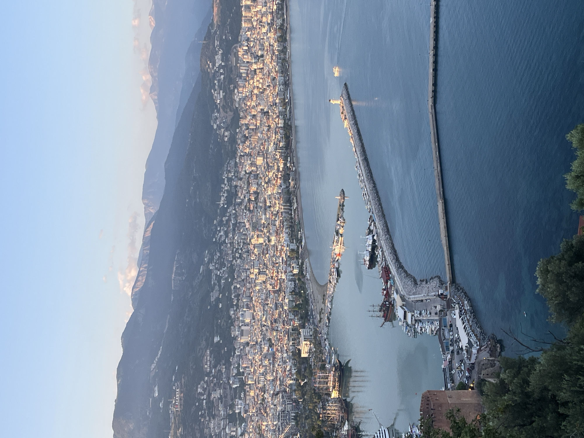 The image shows an aerial view of Alanya, Turkey, captured at what appears to be sunrise or sunset. A mountain range looms in the background, partially obscured by haze. The urban area below is densely populated with buildings that are uniformly colored, stretching along the coastline. A beach runs parallel to the city, with buildings lining it. A long pier extends into the turquoise sea, boats docked along it. A second, longer barrier is visible further out in the water. The lower right corner contains foliage and a partial view of the Alanya Castle.