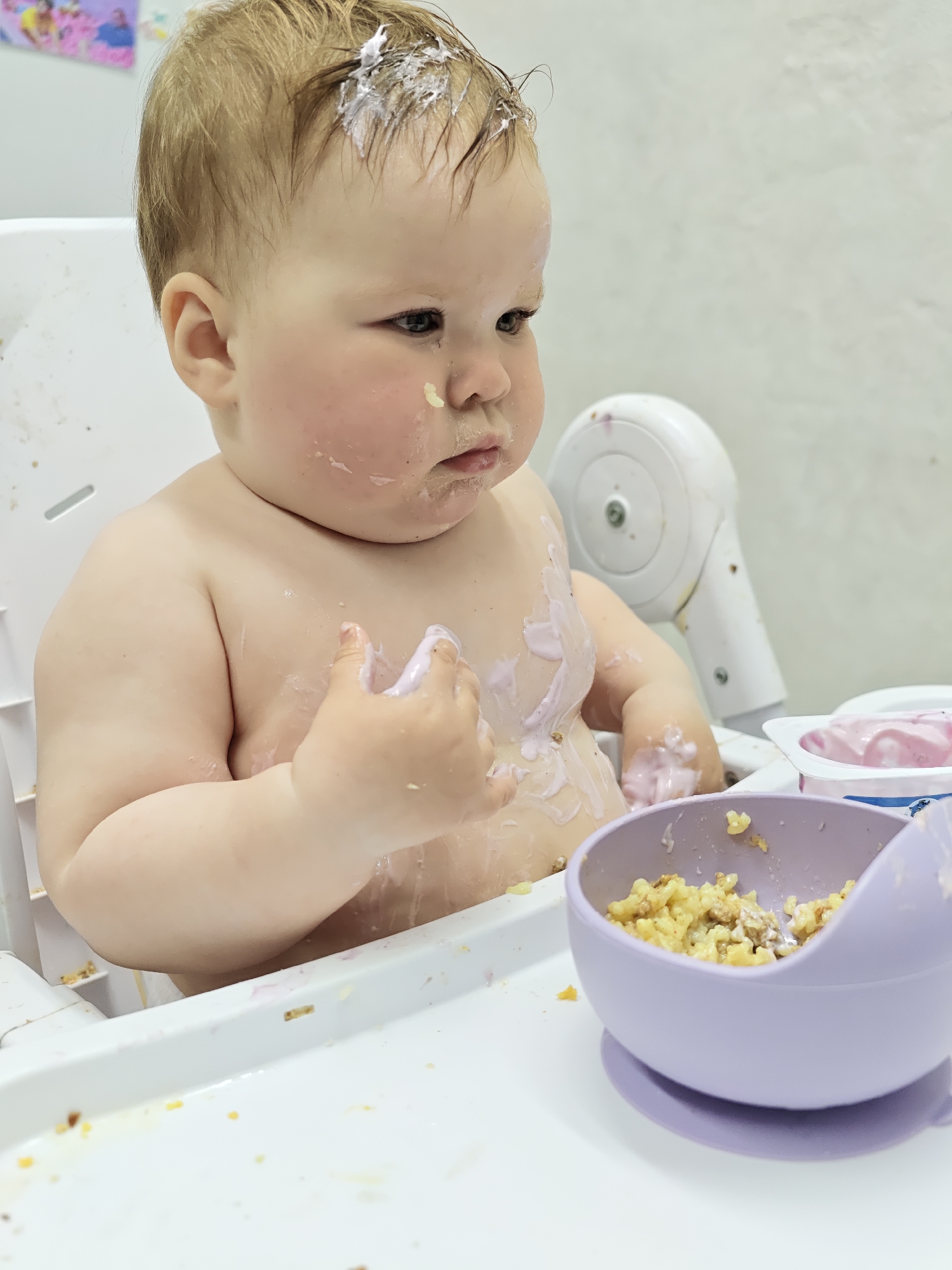 A fair-skinned baby sits in a white highchair, covered in food, seemingly in a domestic setting. The baby has food smeared on their head, face, chest, and hands, and appears to be looking to the side. A purple bowl, containing food that looks like a mix of pasta and meat, sits on the highchair tray. Also on the tray is a container of pink yogurt. The background shows a white wall with some colorful artwork in the top left corner and a white machine attached to the highchair. It is not possible to confirm the location in the city of Chaikovsky, Russia, based solely on the visual information available in the image.