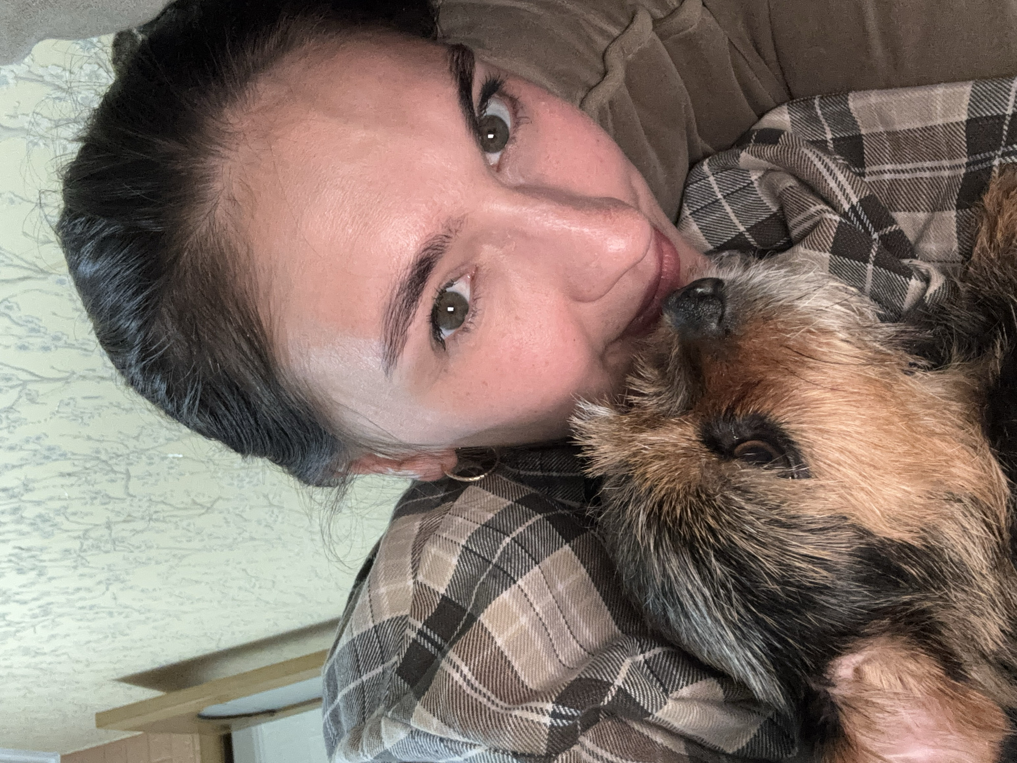 A woman and a dog are lying down. The woman has dark hair pulled back, light skin, and green eyes. She is wearing a brown shirt and a small gold hoop earring is visible in her left ear. The dog is brown and black with wiry fur. They are lying on a brown and gray plaid blanket or shirt. The background is a light-colored wall with a subtle pattern. There is a hint of a light-colored wooden cabinet or frame in the lower left corner. The image is rotated counterclockwise. There is no clear evidence that the photo was taken in Sochi, Russia.