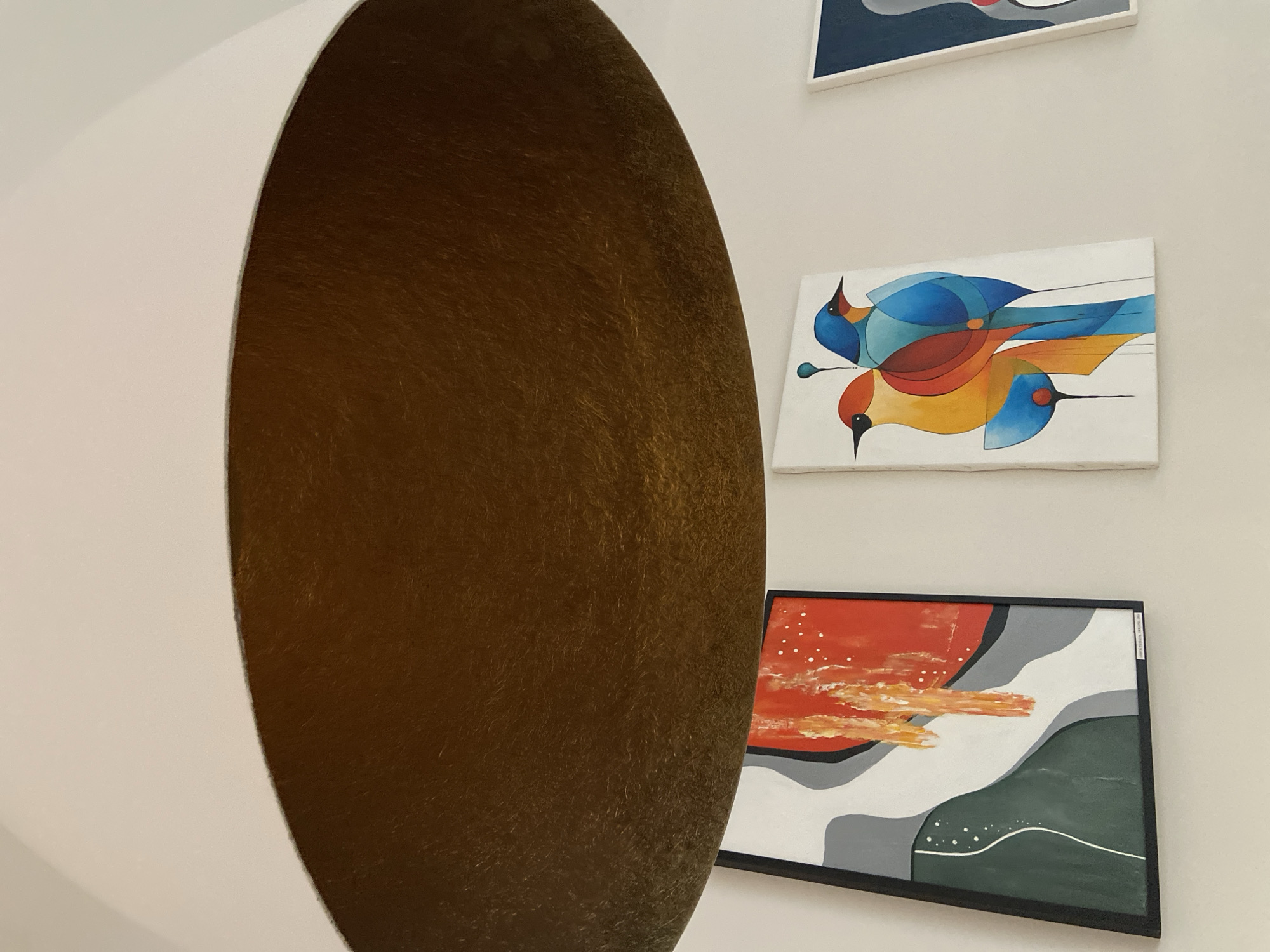An interior scene in Boreti, Montenegro, features a large, off-white, bowl-shaped ceiling light fixture with a textured gold or bronze-colored interior, dominating the upper portion of the frame. Below it, on a clean white wall, three distinct framed artworks are visible. The leftmost artwork is an abstract composition with a black frame, depicting segmented shapes in dark green, light grey, white, and a vibrant red-orange with white specks and brushstrokes. To its right, a second unframed canvas displays an abstract rendering of two stylized birds, one primarily yellow and orange, the other blue, constructed from geometric shapes and lines on a white background. A third, partially visible artwork on the far right appears to be another abstract piece, featuring a grey background with a red circular element. No people or actions are depicted.