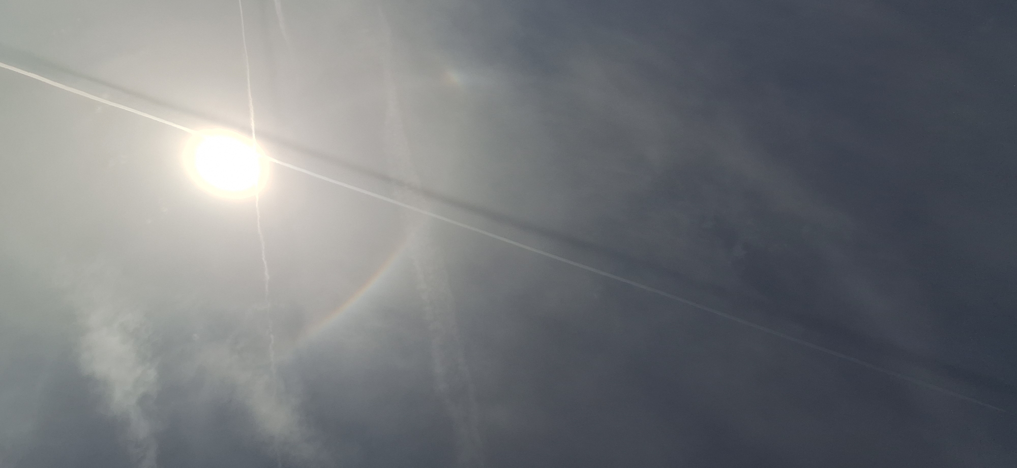 The image shows a bright sun with contrails in a cloudy sky. A faint, partial halo is visible around the sun. The location is not discernible from the image; there is no information to determine if the image was taken in Blagoevgrad, Bulgaria.