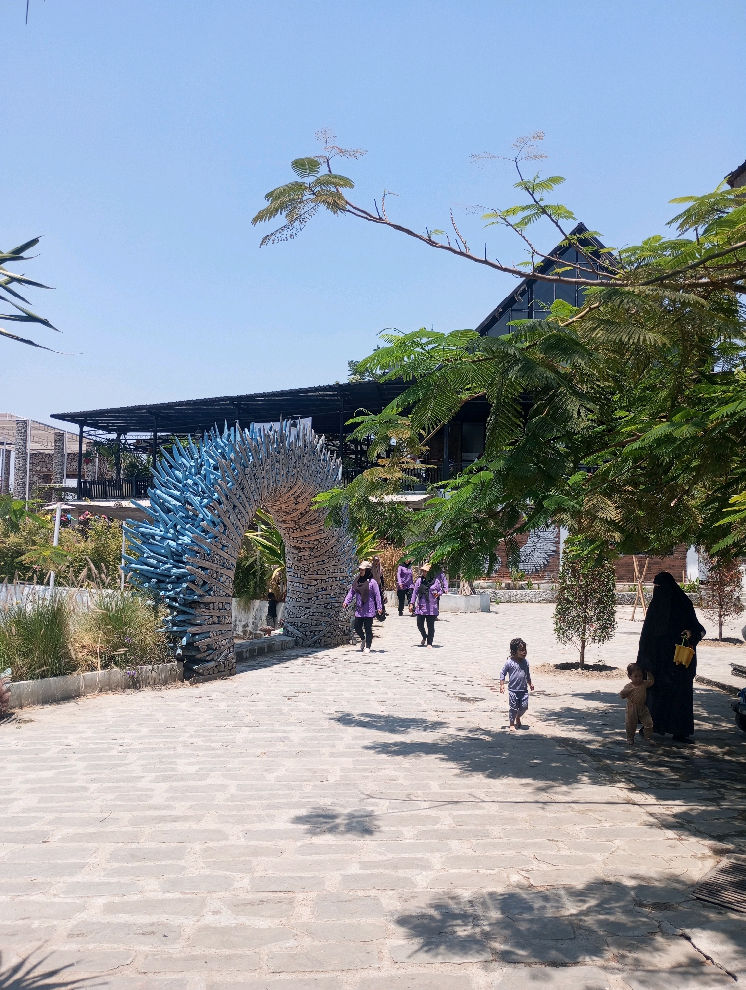 This image captures an outdoor scene in Ponggok, Indonesia, on a bright, sunny day. In the foreground, a paved walkway made of light-colored rectangular stones stretches into the distance, dappled with the shadows of overhead trees. To the left, a large, arch-shaped sculpture constructed from numerous blue and grey wooden elements dominates the view. Beyond the sculpture, lush greenery and vegetation are visible, with various plants and grasses. In the mid-ground, several people are walking along the pathway. Three individuals wearing purple tops and dark bottoms are walking towards the left of the frame. Closer to the right side of the mid-ground, a child in a grey outfit is walking away from the camera, and another child in a tan outfit is walking towards the right. An adult figure in a long black garment, holding a yellow bucket, stands to the right of the children. In the background, a modern building with a dark, sloped roof and large windows is visible, partially obscured by the branches of a large tree with dense green foliage. The sky is clear and blue.
