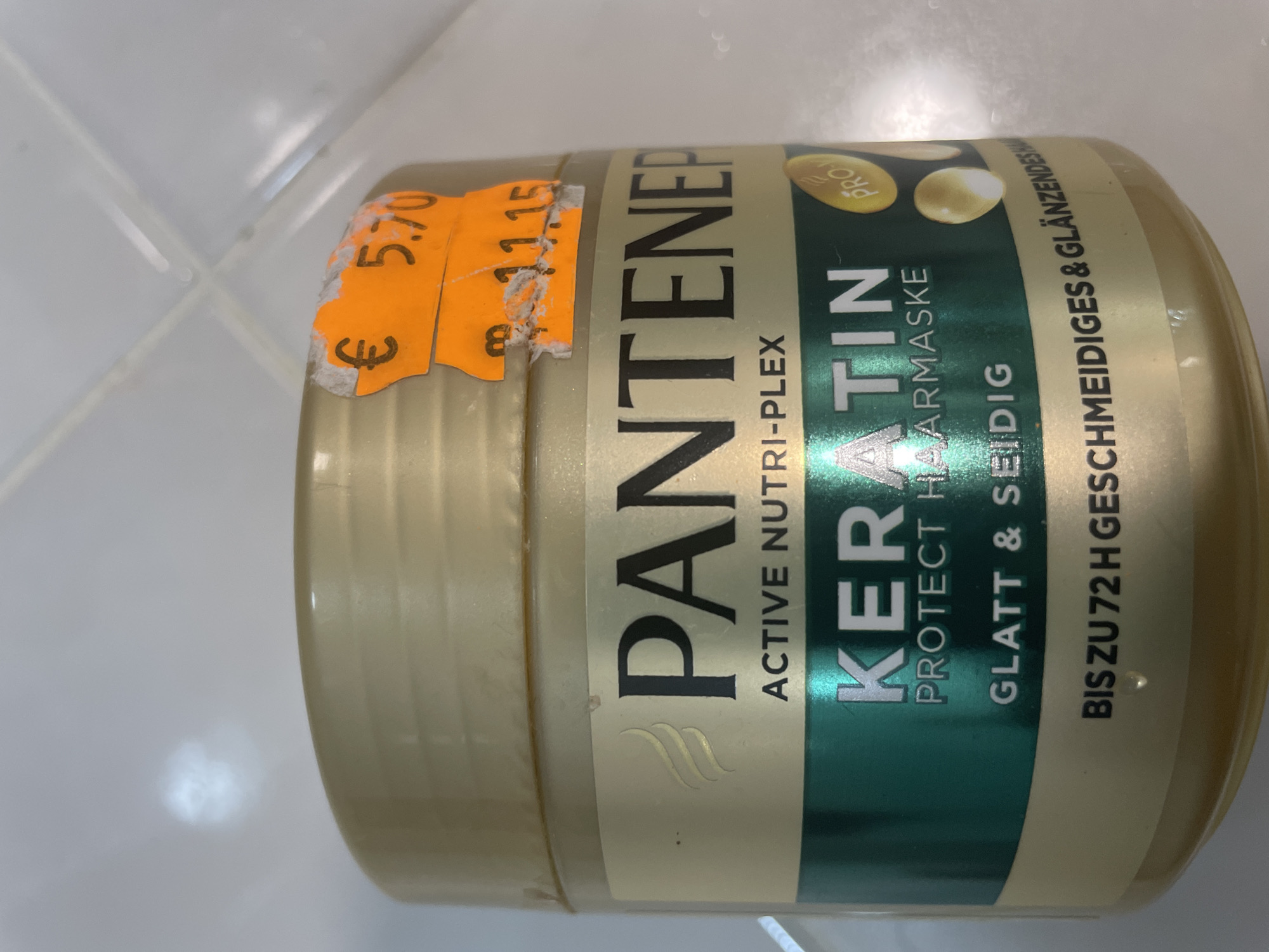 The image shows a close-up of a Pantene Pro-V Keratin Protect Haarmaske hair mask jar. The jar is gold with a green label that reads "KERATIN PROTECT HAARMASKE" and "GLATT & SEIDIG BIS ZU 72H GESCHMEIDIGES & GLÄNZENDES HAAR". There are two price stickers on the jar. One sticker is orange and has "€ 5.70" written on it. Another orange sticker is partially torn and has "B 11.15" written on it. The background appears to be a tiled surface, possibly a bathroom wall. The image does not contain any information about the city of Sevlievo, Bulgaria.