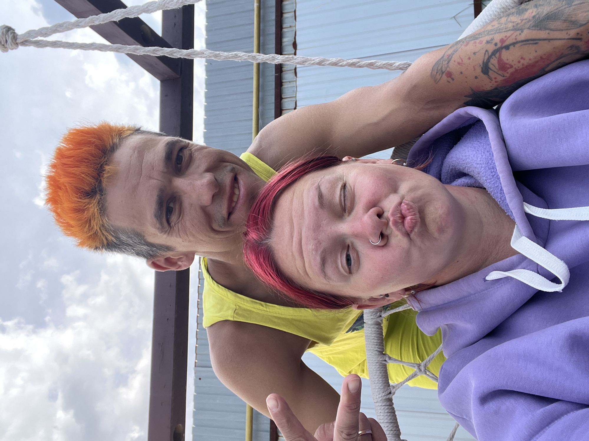 In Chaikovsky, Russia, two individuals are captured in a selfie outdoors. The person in the foreground, a woman with bright red hair and a purple hoodie, is puckering her lips and winking, her nose pierced with a silver ring. Beside her, a man with vibrant orange hair and a yellow tank top places his arm around her. His other hand is raised, with fingers extended in a gesture, and he is smiling at the camera. The background reveals a cloudy sky and a structure that appears to be a wooden frame with ropes, suggesting they are on a swing or a similar outdoor seating arrangement. The overall scene suggests a casual and playful moment shared between the two individuals.
