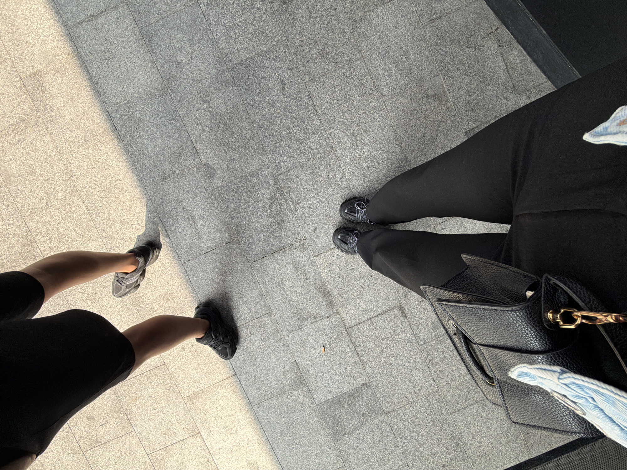 The image shows a view of the ground, revealing two pairs of legs and shoes on a tiled pavement. The tile color varies from light to dark gray due to sunlight and shadow. One pair of legs, on the left, is wearing black shorts or a short skirt and gray sneakers. The other pair is wearing long black pants and dark sneakers, with a black handbag on the right side. The photo appears to be taken in an urban environment, indicated by the tiled pavement. The location is Bayrampaşa, Turkey.