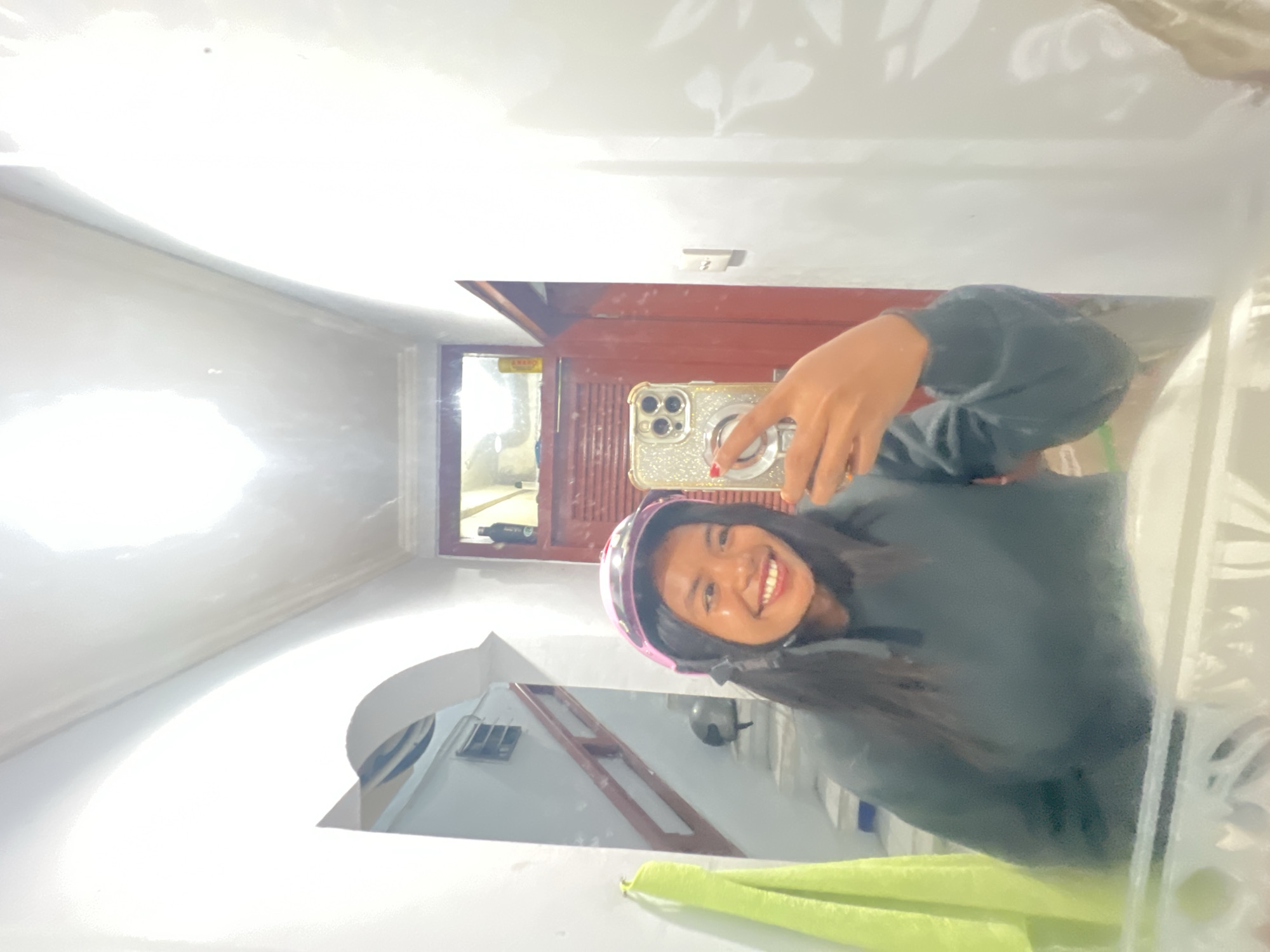A young woman wearing a pink helmet and a dark gray hoodie takes a mirror selfie in what appears to be a hallway with an arched ceiling. She is smiling broadly and holding a gold-encrusted phone in her right hand. In the reflection, stairs with red banisters are visible, along with a lime green towel hanging on the left wall. A second helmet can be seen on the stairs. The environment suggests an interior residential space, possibly in Kuta, Indonesia, indicated by the style of the stairs and railings. The lighting is bright, with a prominent light source above, creating glare on the ceiling.