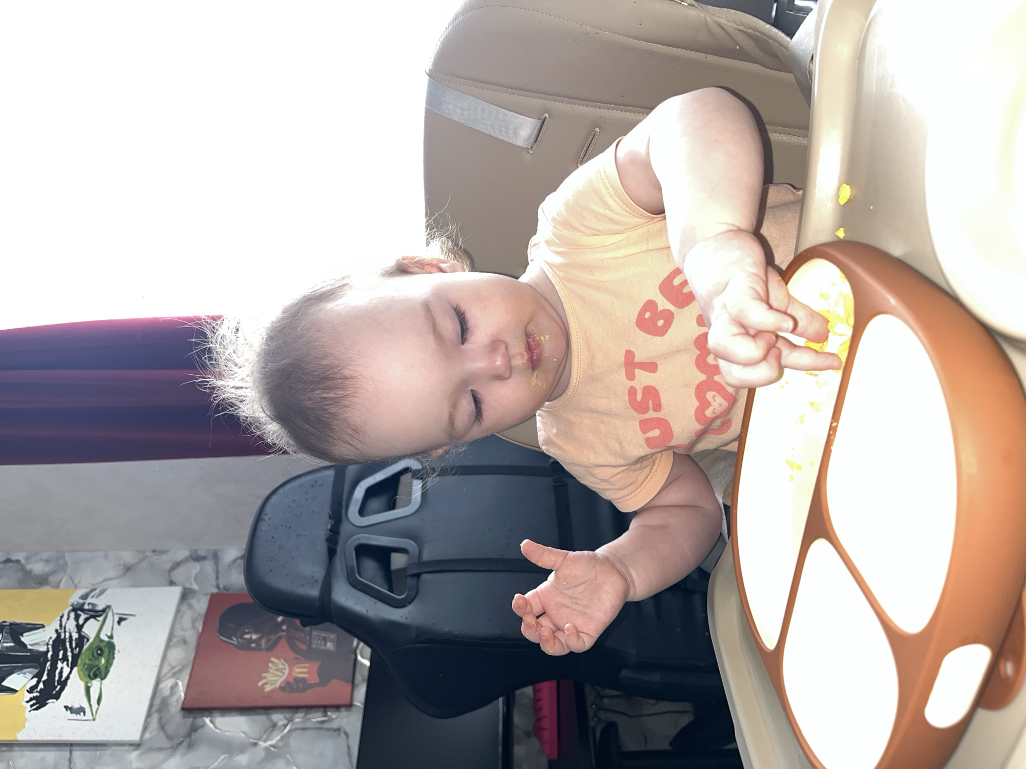 A baby is sitting in a high chair, eating food from a divided plate. The baby has food around their mouth and on their hands. The chair is positioned next to a black gaming chair, and in the background, there are paintings on a wall with a marble-like pattern. A window is visible, letting in sunlight, next to a burgundy curtain. There is no direct evidence indicating the location in the city of Shushary, Russia.