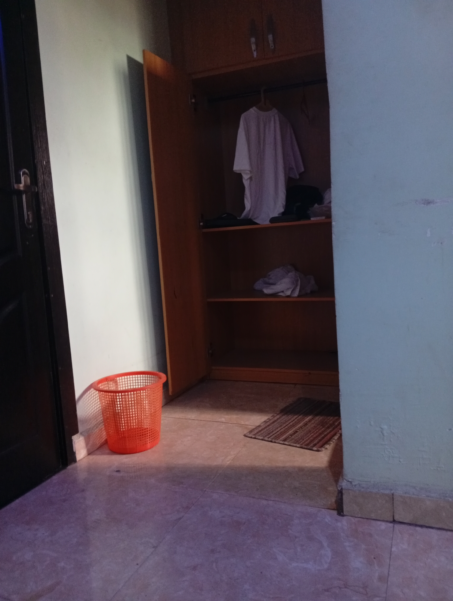 The image displays an indoor scene, likely a room within a residence in Lokoja, Nigeria, characterized by a utilitarian aesthetic. An open, two-door wooden wardrobe dominates the right side of the frame, revealing its contents. A white T-shirt hangs from a hanger on the top rod inside the wardrobe. Below it, the top shelf holds some dark-colored items, while the middle shelf contains a pile of folded light-colored fabric, possibly clothing. The bottom shelf of the wardrobe is empty.
To the left of the wardrobe, against a light grey or off-white wall, stands a bright orange, mesh plastic basket on the tiled floor. A dark wooden door frame with a metal latch mechanism is visible on the far left. The floor is covered with light-colored, possibly beige or subtly purple-hued, square tiles. A small, rectangular doormat with brown and reddish stripes lies on the floor directly in front of the wardrobe's opening.
The room features contrasting wall colors: a light grey or off-white wall on the left and a faded light green or blue-green wall on the right, which shows signs of wear and chipped paint. The lighting appears somewhat dim, casting shadows from the objects, suggesting indoor illumination or a low-light environment. There are no people visible, and no discernible text is present in the image. The scene depicts a functional, lived-in space.