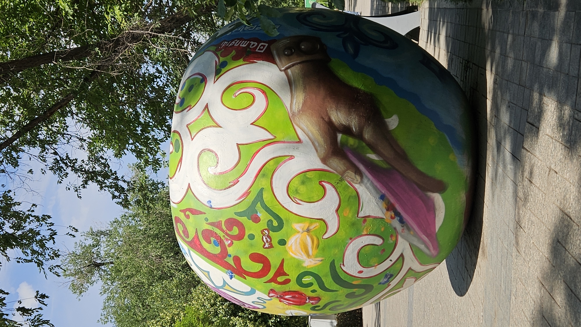 A large, spherical sculpture, painted with a vibrant green base and white, red, and blue decorative patterns, sits outdoors on a paved walkway. The sculpture features a brown, painted hand reaching out, holding a pink object. Various small, colorful shapes resembling candies are scattered across the green surface of the sphere. Trees with green leaves and a blue sky with white clouds form the background. The scene appears to be in a public park or plaza. The sculpture is located in Almaty, Kazakhstan, specifically in the First President's Park.