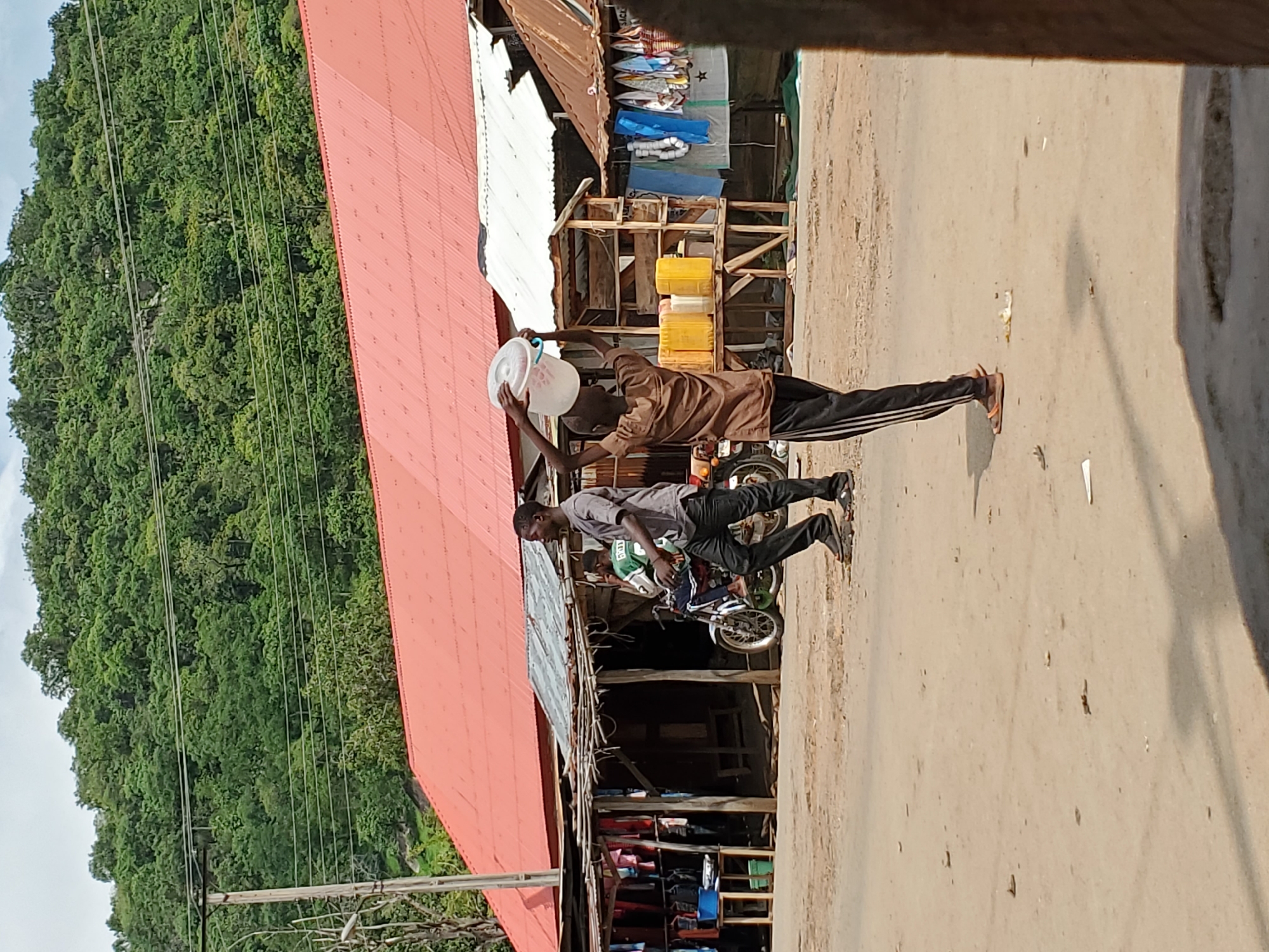 The image shows an outdoor scene on a street in Mararaba, Nigeria. Two men are present. One is standing near a building with a red corrugated iron roof, holding a white bucket above his head. He is wearing a brown shirt and dark pants with white stripes. The other man is seated on a motorcycle facing to the right, wearing a gray shirt and dark pants. The street is paved and appears dusty. Adjacent to the building with the red roof, there are market stalls selling various goods, including blue tarps and clothing. Behind the buildings and stalls, a heavily forested hillside rises. Power lines are visible in the top left of the frame.