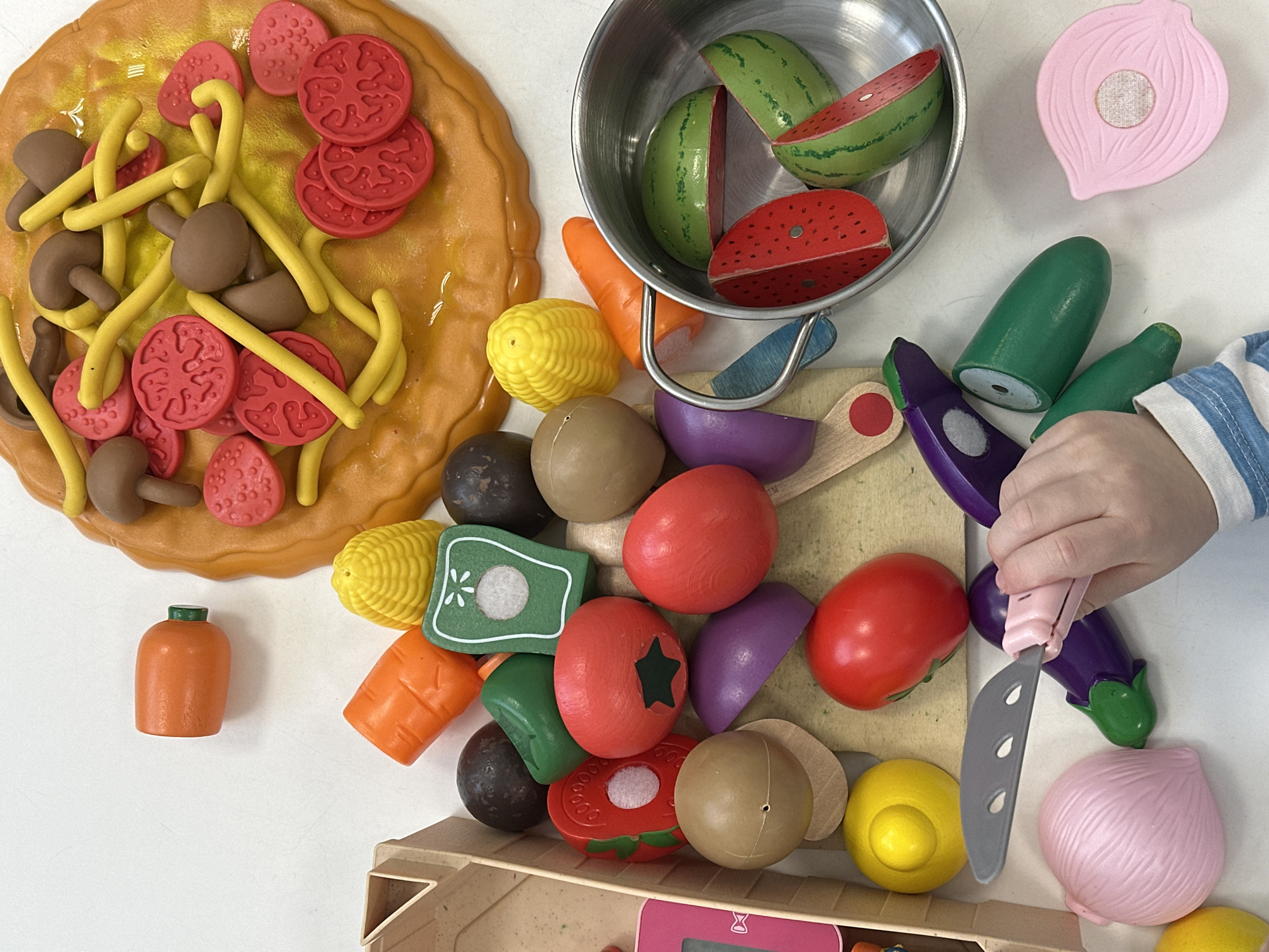 The image depicts a collection of toy food items arranged on a white surface. The food items include a pizza with mushrooms, tomatoes, and pasta, watermelon slices in a metal bowl, corn, carrots, tomatoes, onions, potatoes, eggplants, and other unidentifiable vegetables. A child's hand is visible, holding a toy knife and cutting one of the eggplants. The scene is likely set indoors, possibly in a home or daycare. There is no information to identify the city of Votkinsk, Russia.