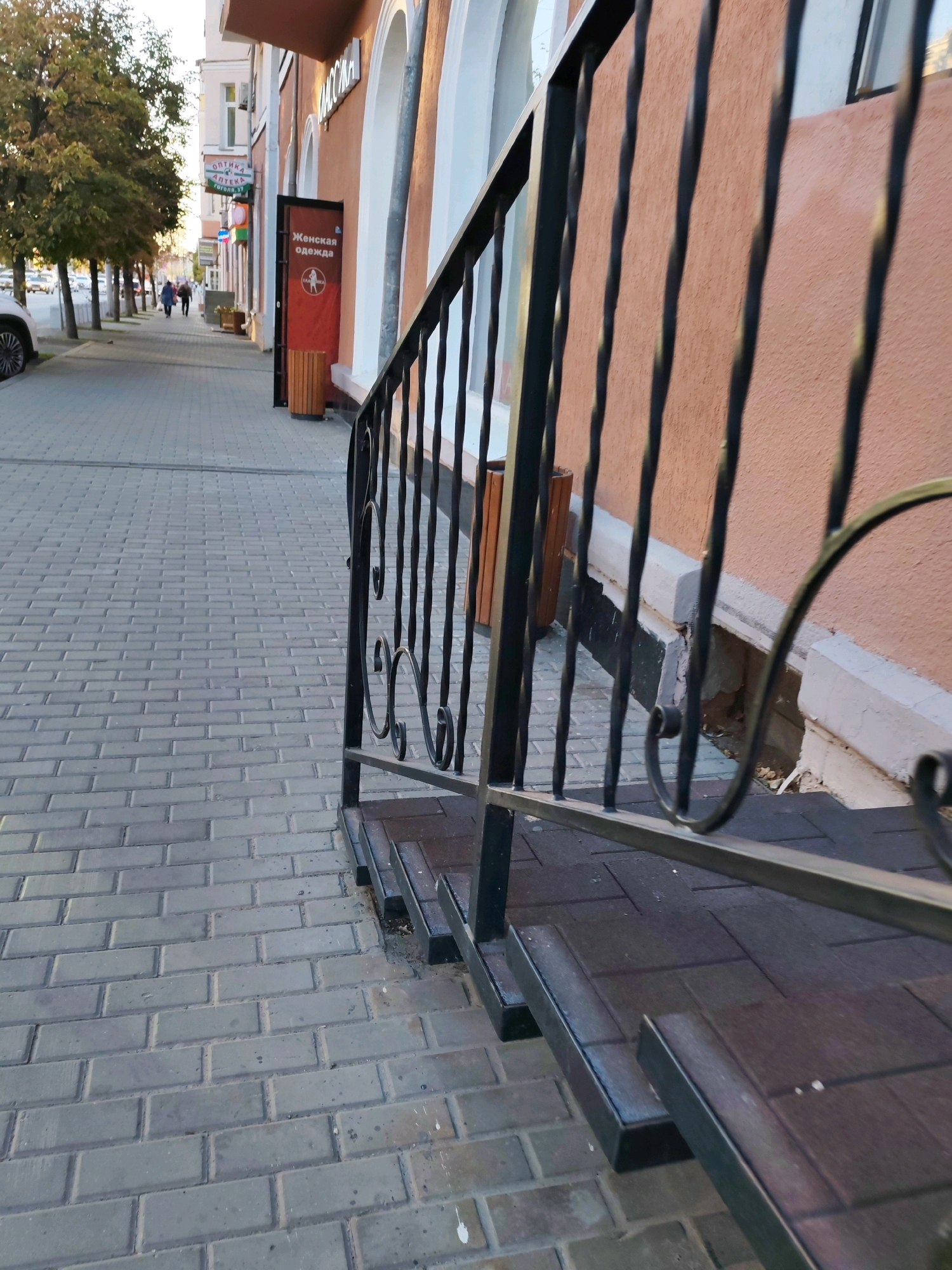 A low-angle, eye-level shot captures a perspective looking down a city sidewalk. The sidewalk is paved with gray rectangular interlocking stones. To the right, a black metal railing with twisted spindles runs along a set of dark-tiled steps. Beyond the railing, a light brown building with a textured facade rises. A bright orange sign with Russian text and a silhouette of a woman in a dress is affixed to the building. Further down the sidewalk, several trees with green and yellow foliage line the left side of the path. Two figures, a woman and a man, walk away from the camera down the sidewalk. A white car is parked on the left, and more buildings and trees are visible in the distance. The scene is illuminated by natural light, suggesting it is daytime.