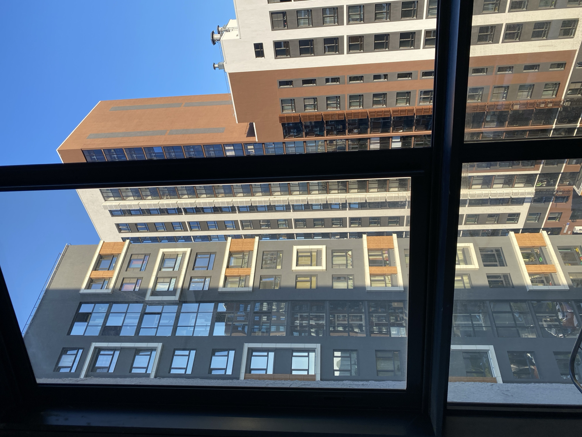 The image, taken from an interior perspective looking out through a window frame, shows a cluster of modern, multi-story apartment buildings against a clear blue sky. The buildings exhibit a variety of architectural styles and color palettes, including grey, white, and terracotta facades. Many of the buildings feature large, reflective windows, some with balconies. The sunlight casts distinct shadows on the structures, indicating it is likely daytime. Reflections of the buildings and the sky are visible on the windows. The scene is devoid of any human presence. The architectural style and the context of modern residential complexes are consistent with urban development found in Yekaterinburg, Russia, particularly in newer residential areas.