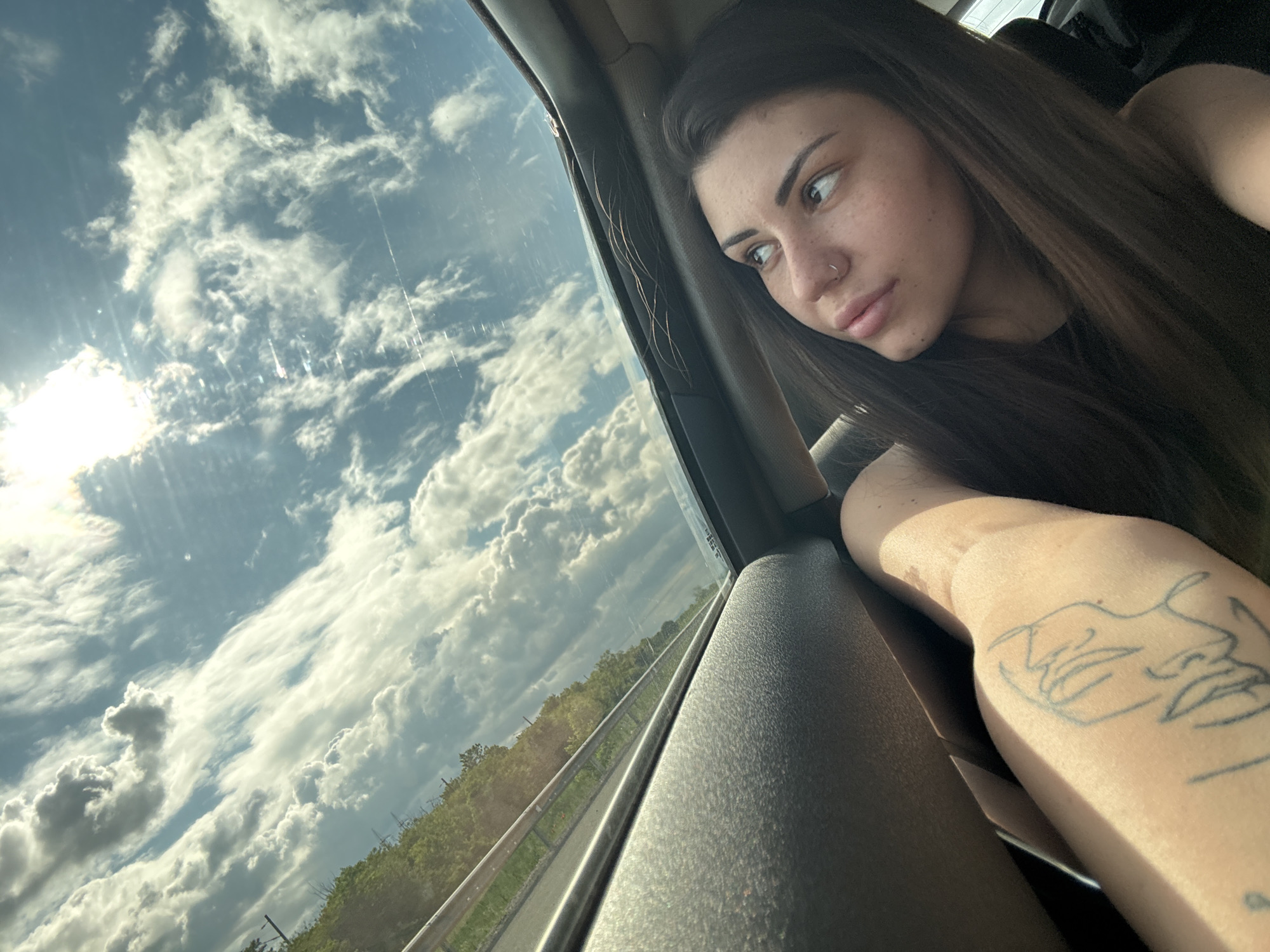 A young adult woman is captured in a close-up shot inside a vehicle, looking out a side window. She has long, dark hair, a nose ring, and subtle freckles on her face, which is turned to profile as she gazes pensively towards the outside view. Her right arm, adorned with a line-art tattoo of two faces, rests on the car's interior panel, bathed in sunlight.
The scene outside the window reveals a vibrant, cloudy sky. Bright white cumulus clouds dominate the blue expanse, with the sun visible in the upper left, casting a strong glare and lens flares across the windowpane, indicating a bright daytime setting. Below the sky, a lush green landscape of trees and foliage stretches along what appears to be a road or highway, partially visible with a metal barrier. The diagonal orientation of the horizon suggests either the car is moving, or the photo was taken with a tilted perspective. The overall atmosphere conveys a sense of quiet observation or contemplation during a journey. The location context is Hrabarsko, Bulgaria. No discernible text is visible other than the artistic tattoo.