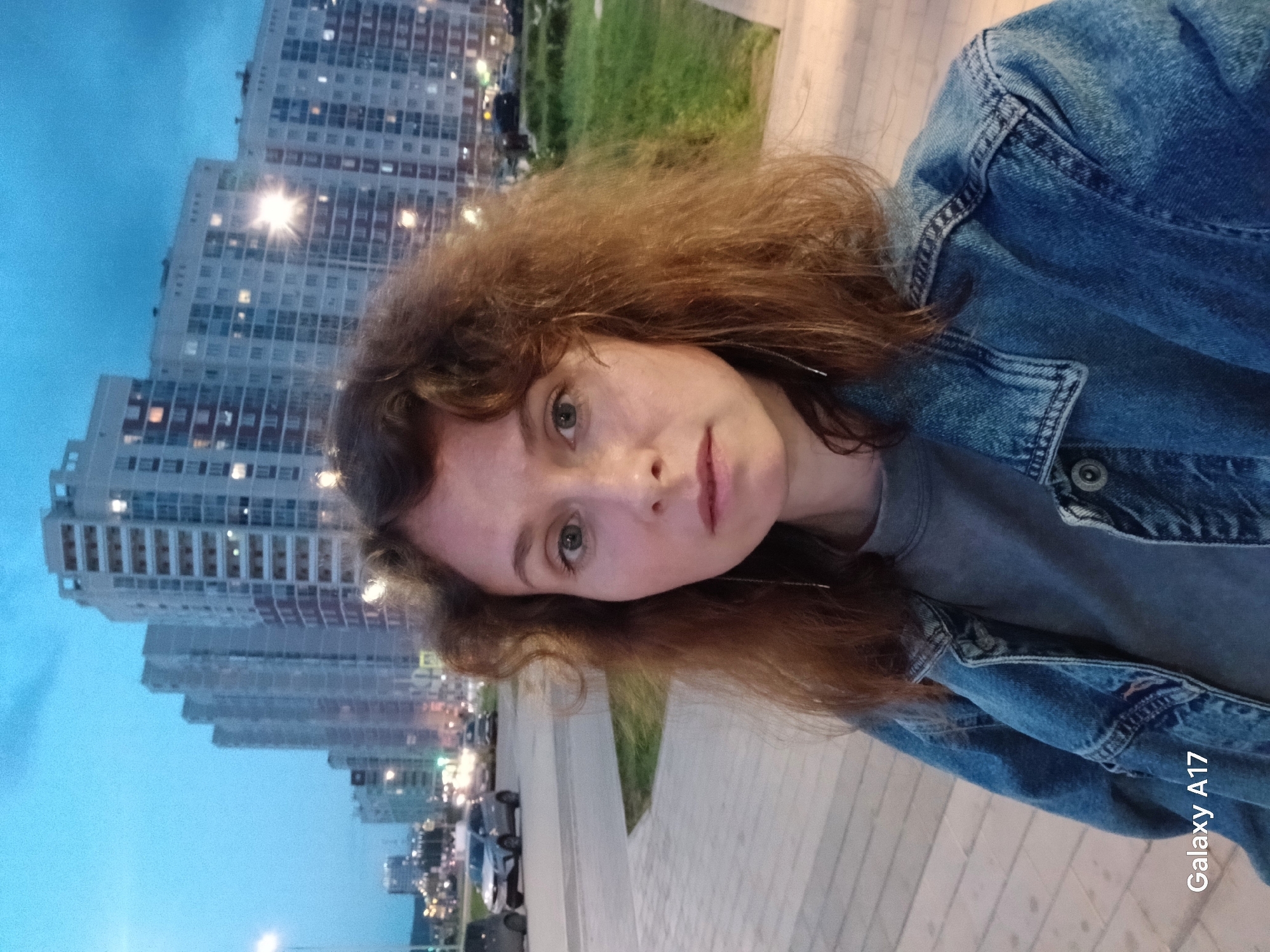 A woman with curly brown hair wearing a blue denim jacket takes a selfie outdoors at dusk. In the background are tall, multi-story apartment buildings, and a street with cars. The photo was likely taken in Yekaterinburg, Russia.