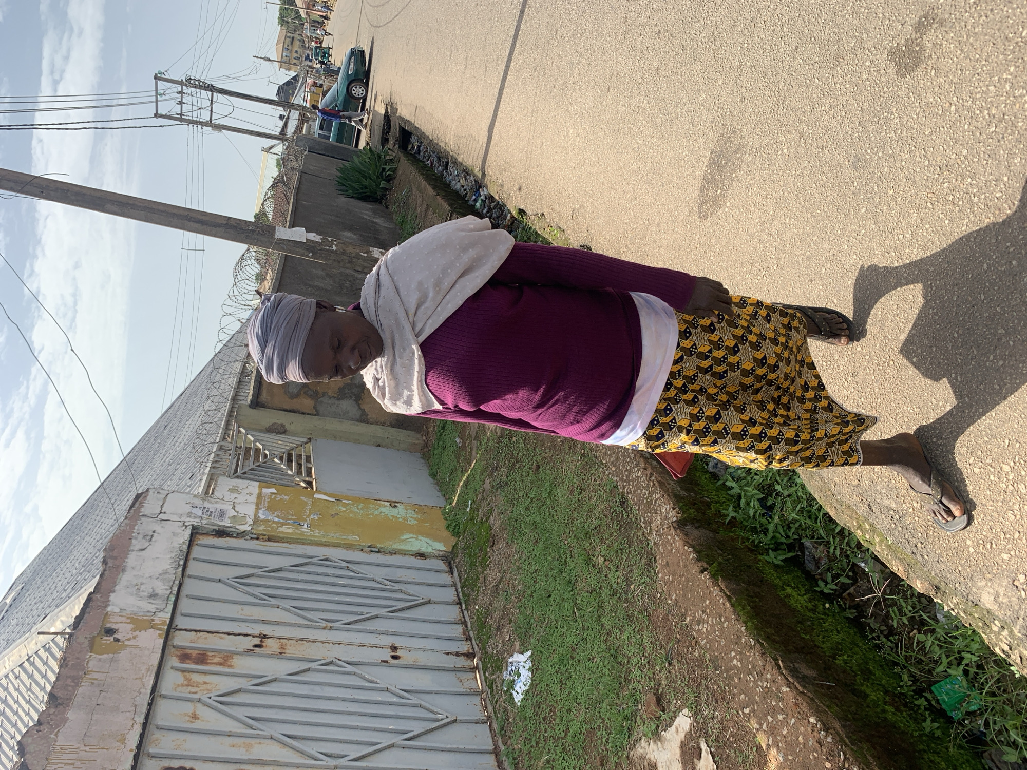 In Jos, Nigeria, an elderly woman wearing a purple long-sleeved shirt, a white undershirt, a patterned yellow and black skirt, and a grey headwrap walks along a paved road. She is also wearing a light-colored scarf draped around her shoulders and dark flip-flops. The woman is walking on a sidewalk that borders a drainage ditch filled with vegetation and debris. To her left, a weathered metal gate and a building with a corrugated metal roof are visible. Behind the woman, a utility pole with numerous wires leans precariously over the road. The road is lined with buildings, fences topped with barbed wire, and other utility poles. A green sedan and a tricycle are also visible on the road in the background. The sky is partly cloudy.