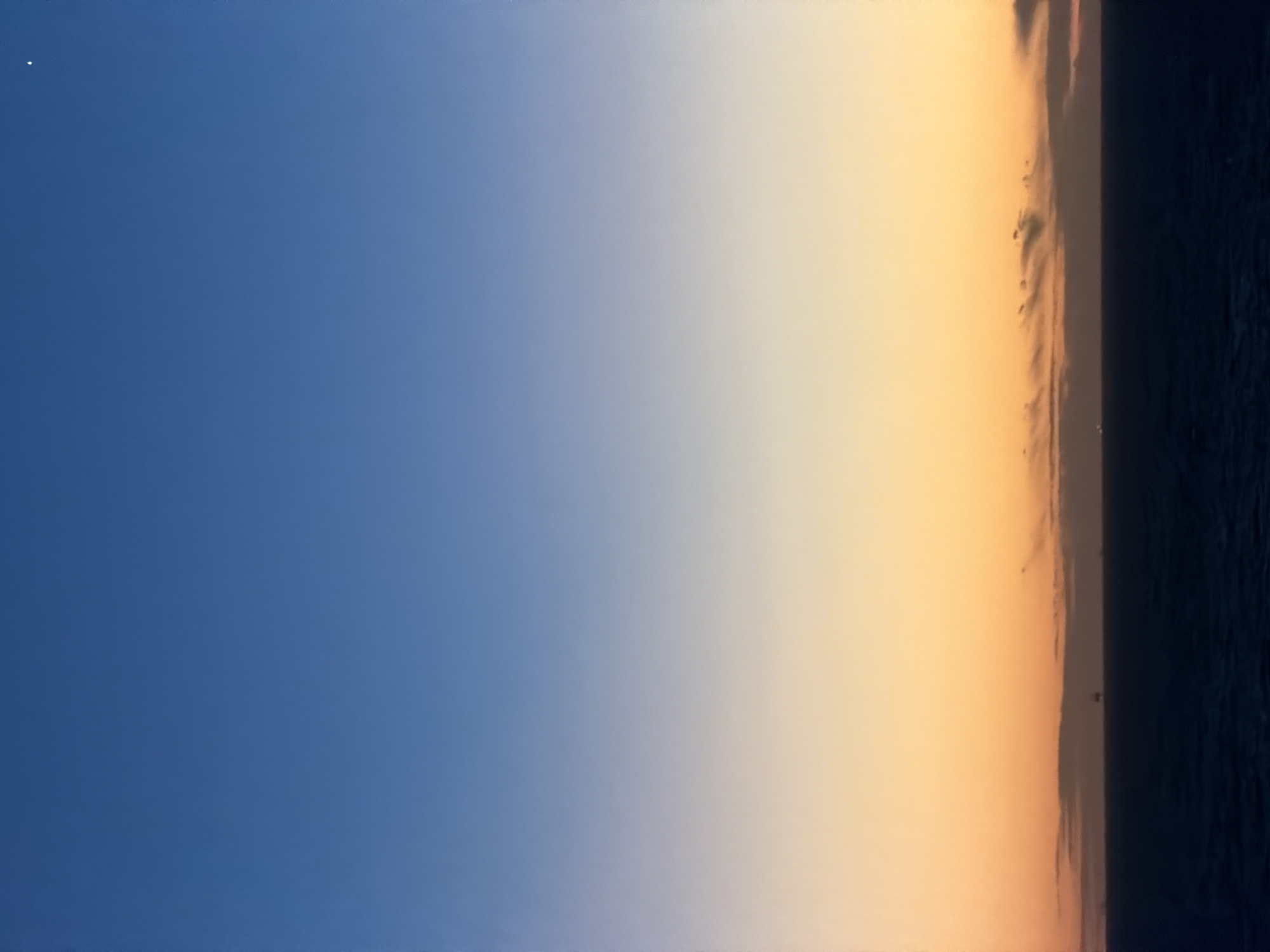 The image depicts a sky transitioning from blue to orange at sunset, with a dark mass, likely water or land, at the right edge. A single, bright dot is visible in the blue portion of the sky, possibly a star or planet. The scene lacks identifiable features to pinpoint its location within any city or region. The scene evokes calmness and the transition of day into night.