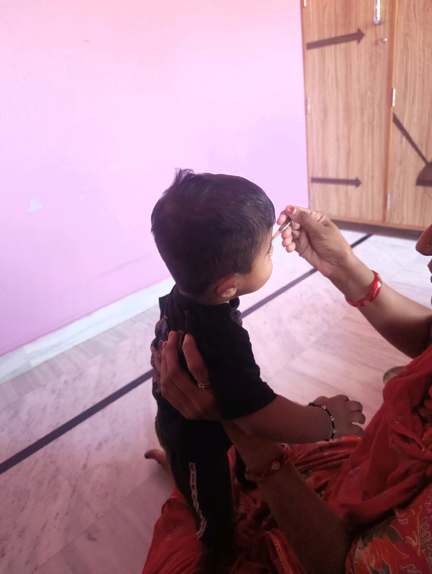 In Chhapri Bari, India, a child in a black shirt is being given a medicinal application to their eye by an adult. The child is facing away from the camera, with their back towards the viewer. The adult, wearing a red garment, is positioned to the right of the child, their hand applying the substance. The environment appears to be indoors, with a pink wall on the left and wooden doors with arrow carvings on the right. The floor is tiled with a dark line dividing sections. The lighting is soft, casting shadows and suggesting a natural light source.