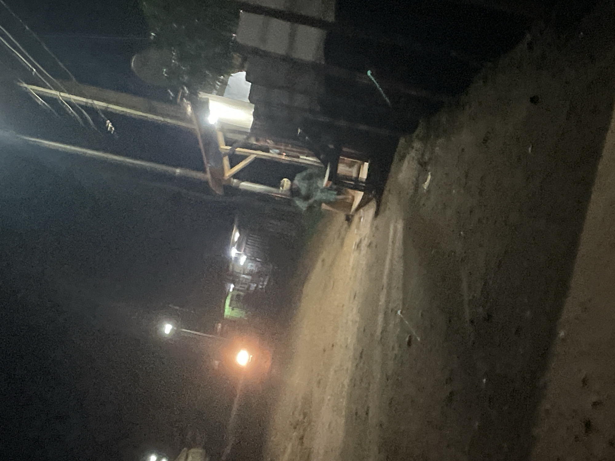 The image depicts a nighttime scene on a dirt road in Lokoja, Nigeria. A single individual, wearing a light-colored garment and head covering, stands near a wooden structure that appears to be a small shop or stall. The structure is illuminated by artificial lighting, and a satellite dish is visible on top of it. Overhead, power lines crisscross the sky, supported by wooden poles. In the background, another light source, possibly a streetlamp or vehicle headlight, casts a warm glow on the scene. The ground is uneven and covered with dirt and debris.