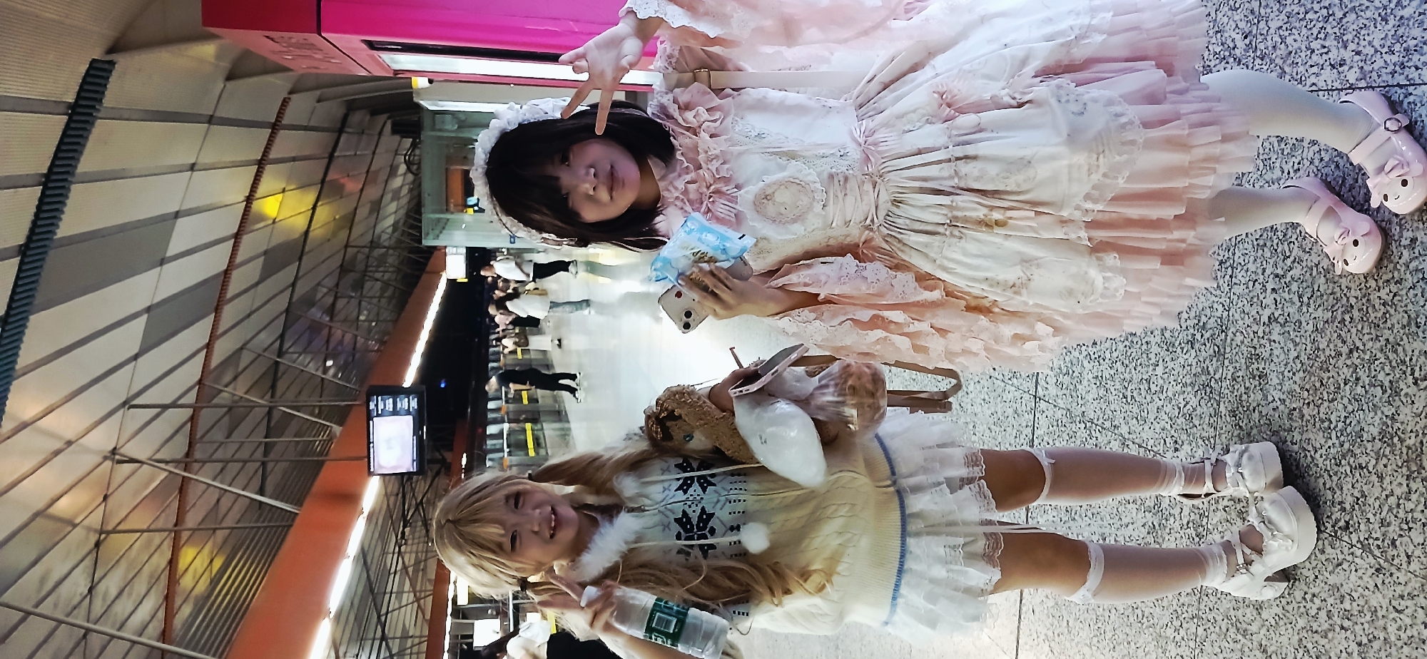 Two young women are posing for a photograph in a subway station in the Chongqing High-tech Zone, China. The woman on top has dark hair, is wearing a white and pink Lolita-style dress with lace and frills, and is holding a smartphone and a package of tissues. She is making a "V" sign with her fingers. She is also wearing pink shoes. The woman below has blonde hair and is wearing a white sweater with snowflakes and a white skirt with frills. She is holding a water bottle and is also making a "V" sign with her fingers. She is wearing white shoes. They are standing on a patterned floor, and there are other people waiting behind a glass barrier in the background. A digital display screen is visible on the left side of the frame.