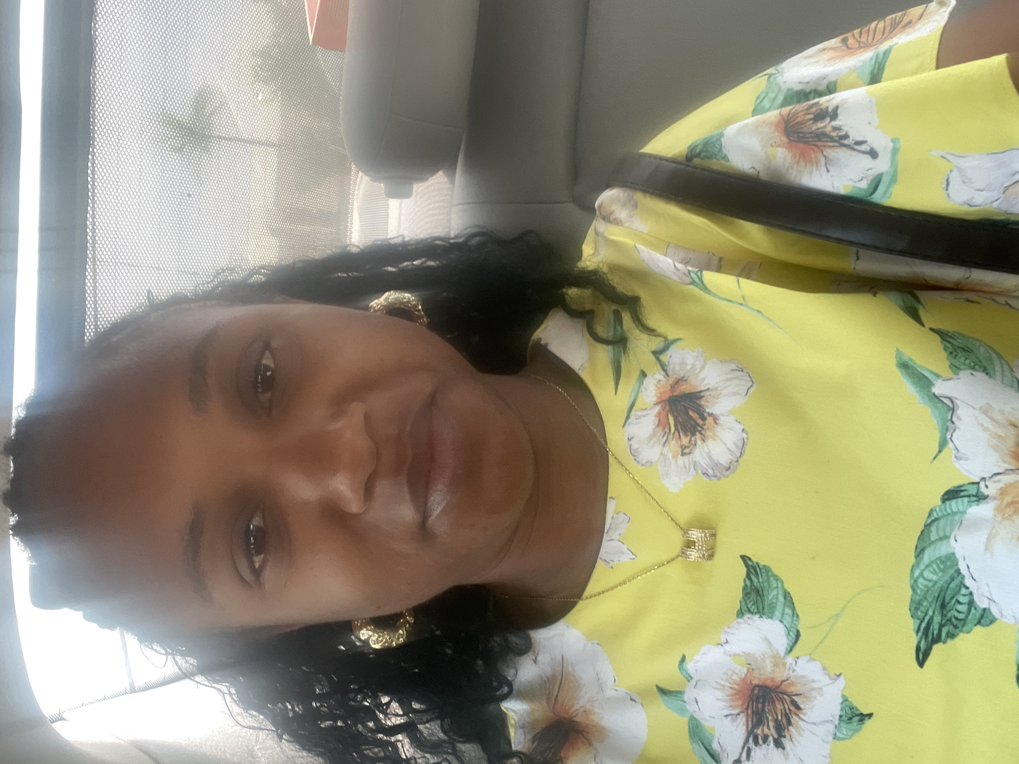 A woman is pictured indoors, possibly in a car, given the presence of a headrest in the background. She is wearing a bright yellow floral print top and gold jewelry, including hoop earrings and a necklace. The background shows a window with a mesh screen through which a palm tree is visible. No information is provided to confirm her location in Jos, Nigeria.