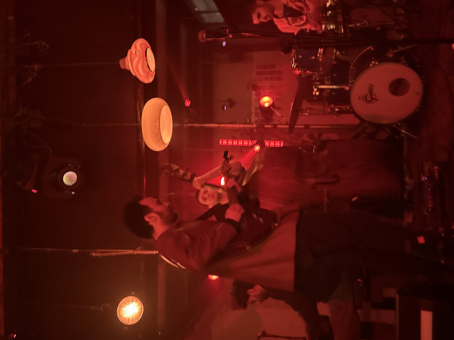 Four male musicians are depicted performing on a stage under dominant red lighting. From left to right: a partially visible male individual with dark hair and beard operates a keyboard; a male individual with dark hair and beard, wearing a dark jacket, plays an electric guitar, positioned in the foreground; another male individual with short dark hair plays an upright bass; and a partially visible male individual with dark hair and beard, wearing a patterned shirt, is seated behind a drum kit featuring a white Gretsch bass drum. The ceiling shows exposed pipes and contains multiple lighting fixtures, including two spotlights, one industrial-style hanging lamp with an exposed bulb, and two pendant lamps with light-colored shades. Thin red light beams traverse the upper midground. The specific location within Tel-Aviv, Israel, is not identifiable from the provided image.