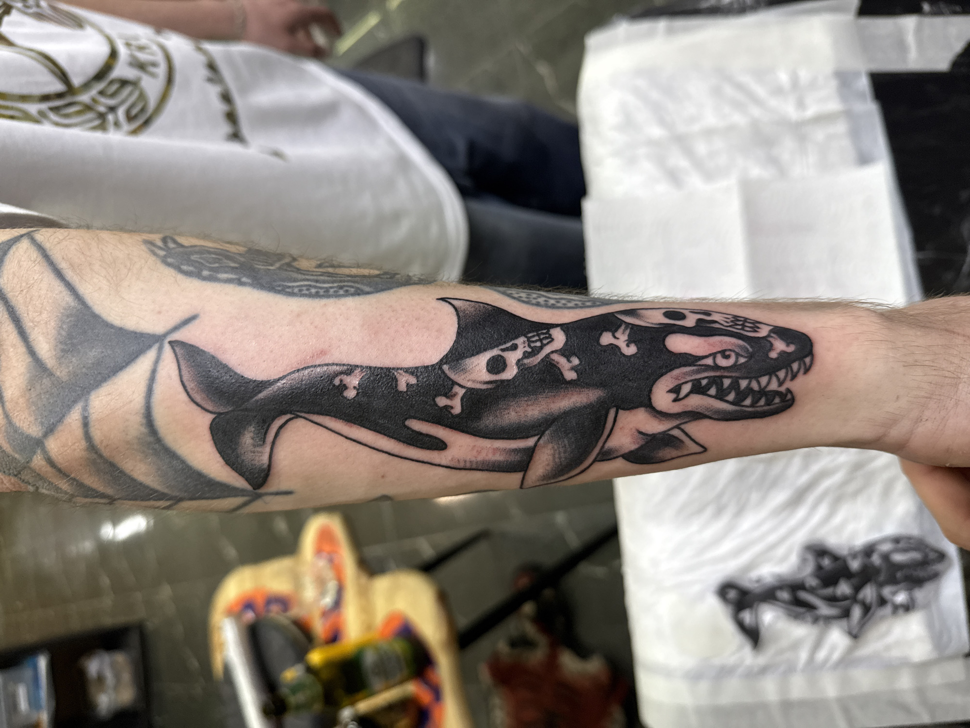 A close-up shot depicts a forearm tattoo of an orca-like creature with skull and crossbones imagery on a person's left arm. The tattoo is primarily black and white. The person's arm is extended, and other tattoos are partially visible in the background. A white shirt is visible, as well as a reflective surface with blurred objects. There is no information about the location of Moscow, Russia.