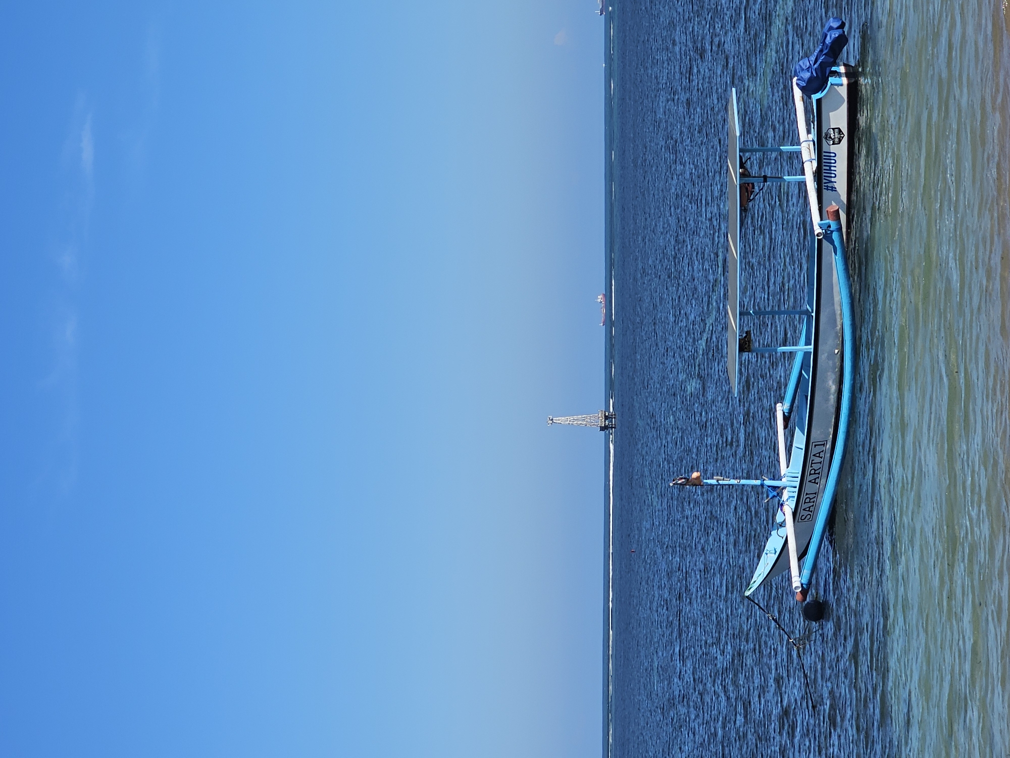 The media file depicts a tranquil scene at sea, with a clear blue sky dominating the left half of the frame and the deep blue ocean stretching across the right. The sun appears to be high in the sky, suggesting a daytime setting, likely mid-morning or early afternoon. The water is calm, with gentle ripples indicating minimal wind.
The primary object of interest is a traditional Indonesian fishing boat, known as a jukung, anchored close to the shore. The boat is painted in vibrant blue and white, with outriggers extending outwards for stability. On the side of the boat, the text "SARI ARTA1" is clearly visible in bold lettering. Towards the stern of the boat, another inscription reads "#YUHUU". A blue tarp is neatly folded and secured at the bow.
In the distance, several ships are visible on the horizon, hinting at a busy shipping lane. Closer to the shore, a solitary man is seated on the boat, possibly a fisherman or someone enjoying the solitude. Further out at sea, a tall, lattice-like structure, possibly a navigation marker or a small offshore platform, stands prominently against the horizon. The water near the shore is shallow and clear, revealing the sandy seabed below. The overall atmosphere is serene and peaceful, conveying a sense of calm and relaxation.