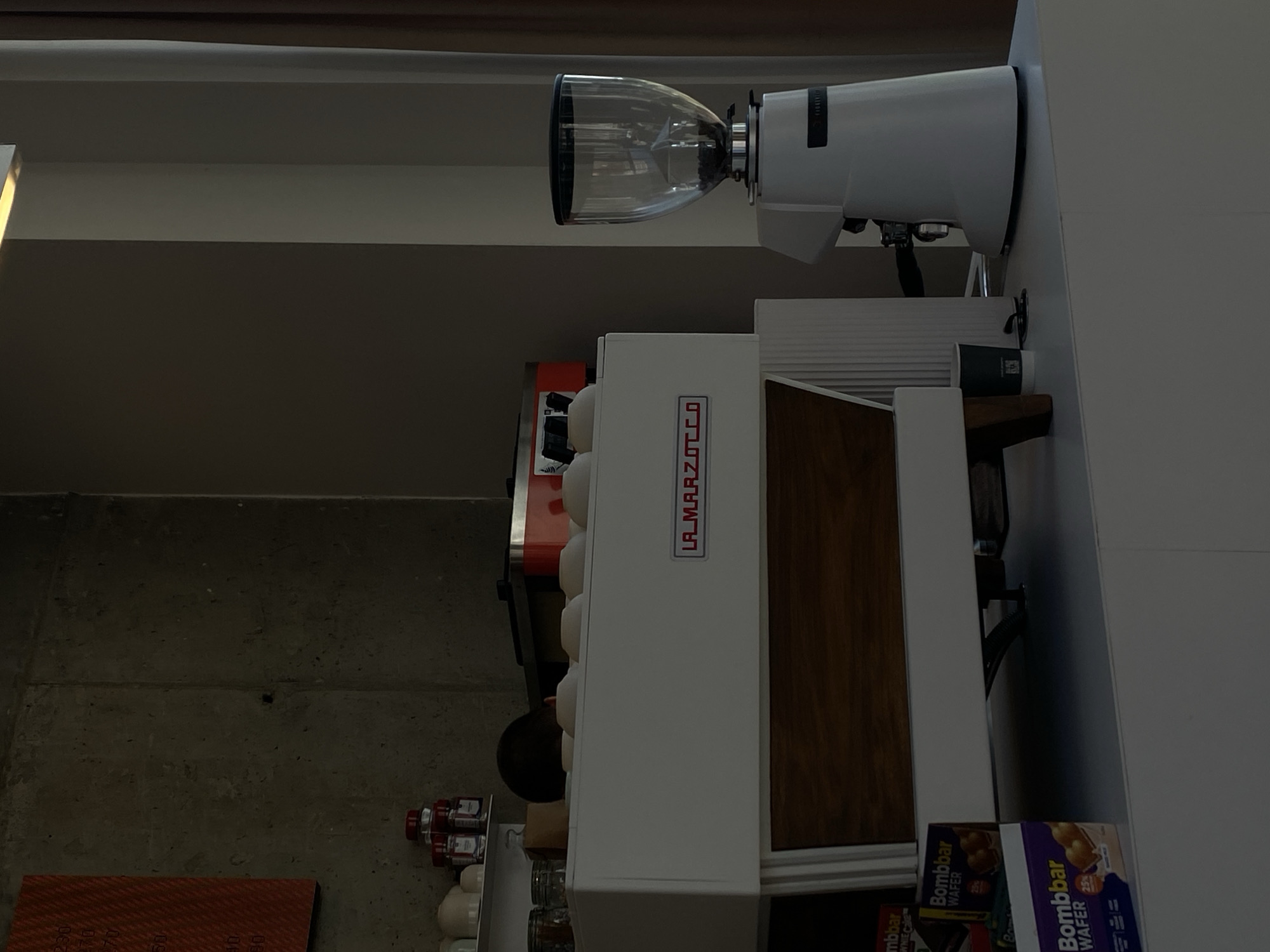 The image depicts a coffee station in a dimly lit interior, possibly a cafe or a home kitchen. Prominently featured is a white La Marzocco espresso machine, with a wooden panel detail on its front. To its right stands a white coffee grinder with a clear hopper filled with coffee beans. In the foreground, to the left of the espresso machine, are two boxes of "Bombbar Wafer" snacks. The background consists of a textured concrete wall on the left and a lighter-colored wall with beige curtains on the right. The overall atmosphere is quiet and still, suggesting a moment of pause. The location of Dinskaya, Russia, is not discernible from the visual information provided in the image.