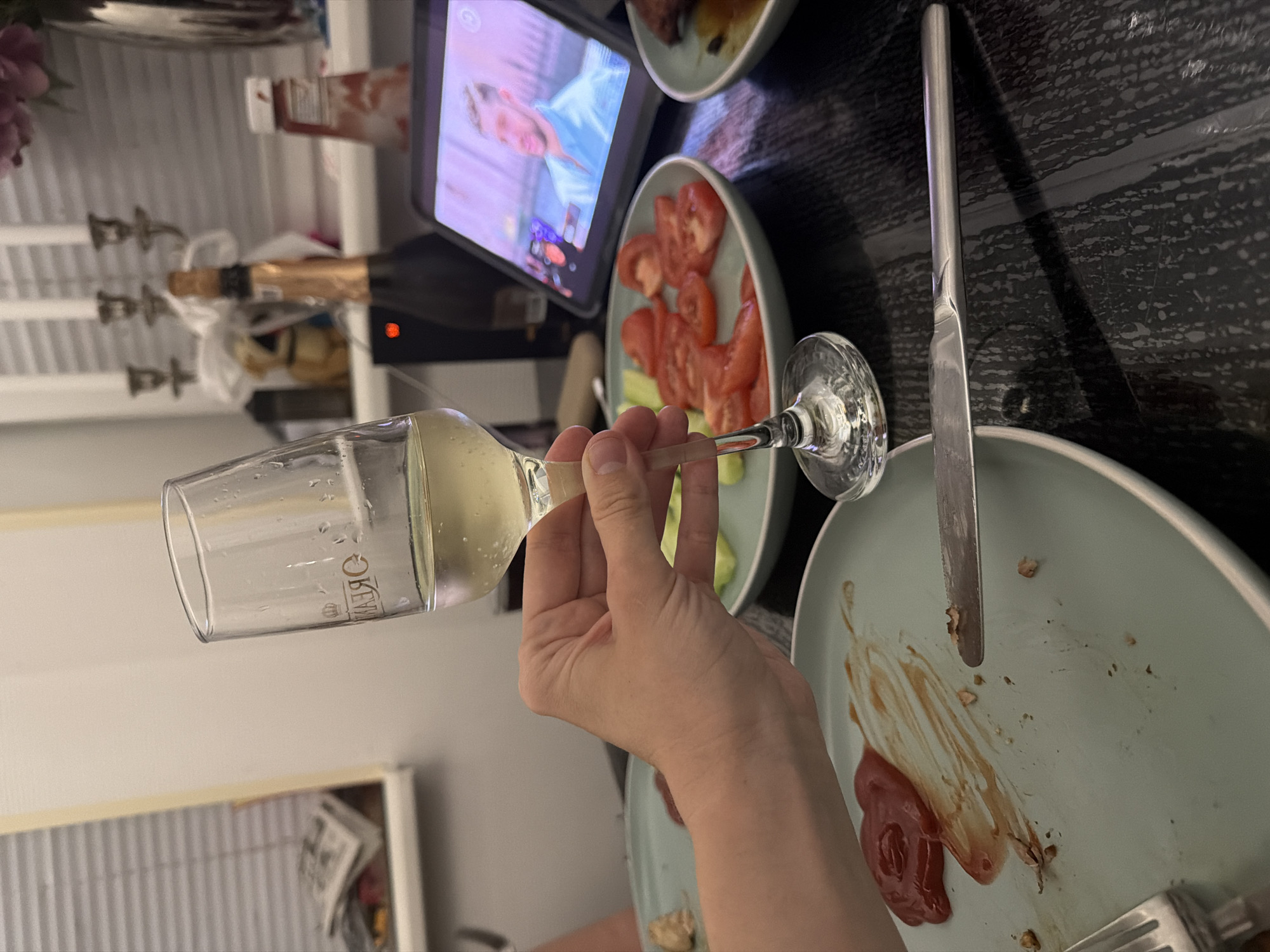 The image shows a close-up of a hand holding a champagne flute filled with a pale yellow liquid. The hand is positioned in the foreground, with the glass tilted. In the background, a plate with sliced tomatoes and cucumbers is visible, as well as a partially eaten meal on another plate with ketchup and some crumbs. A bottle of champagne and a tablet displaying a video of a man are also present on the table. The setting appears to be a dining table with a dark, textured surface. The lighting suggests an indoor environment, possibly a home or restaurant. The overall scene depicts a meal or celebration in progress. The location in Bila Tserkva, Ukraine is not discernible from the visual information provided.