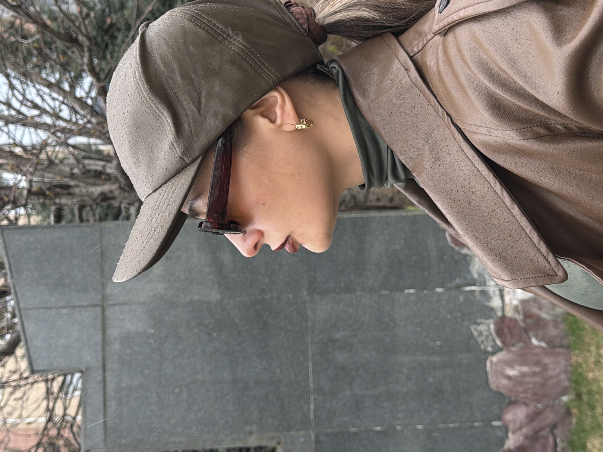 A person wearing a khaki baseball cap, sunglasses, and a brown leather jacket is seen in profile. The cap and jacket are covered with raindrops. The person has a gold earring visible on their left ear. They are also wearing a green turtleneck shirt. The background includes a grey tiled ground, bare tree branches, and some greenery.
The image provides no evidence of the location being in Krasnoyarsk, Russia.