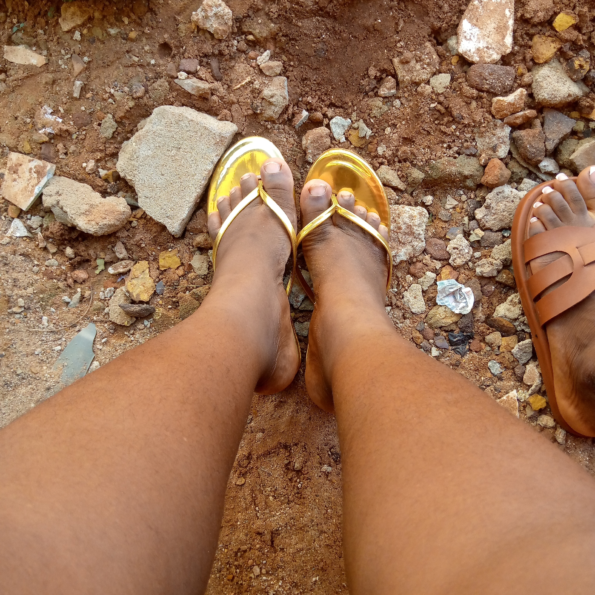 The image shows a first-person perspective of two bare legs standing on a surface of reddish-brown earth mixed with small rocks and debris. The left foot is wearing a gold-colored sandal with a thong design, while the right foot is wearing a brown sandal with a cross-strap design. A small piece of crumpled silver foil lies between the sandals. The scene is in Ukwu-Abwa, Nigeria.