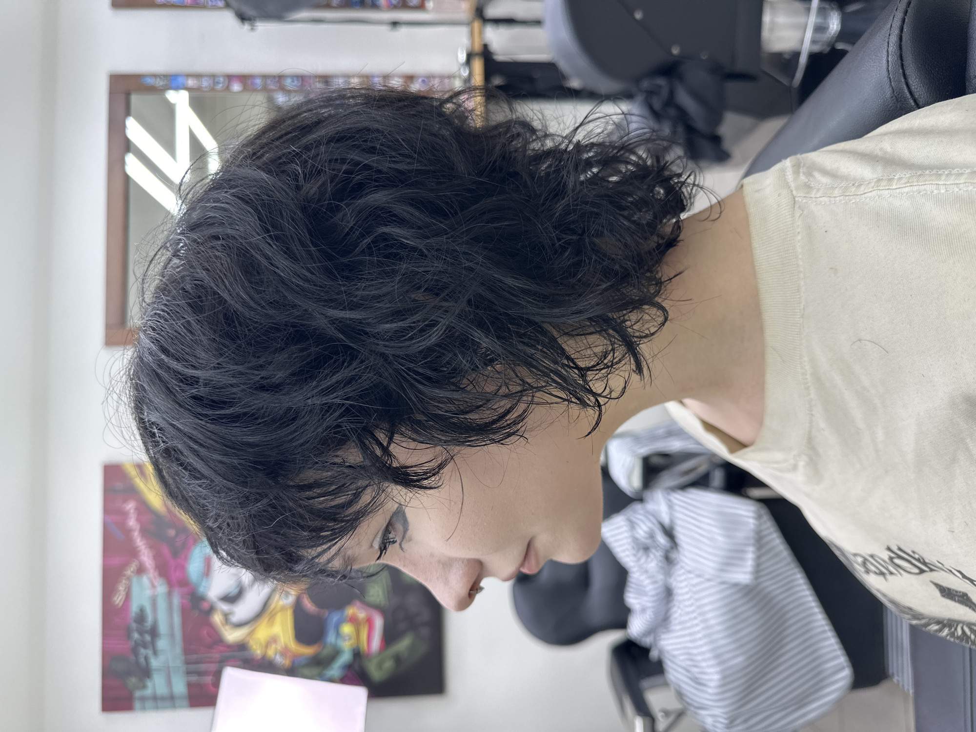 A medium shot captures a person from the side view, likely inside a salon in a location unidentifiable as Chaikovsky, Russia. The individual has short, dark, wet hair styled in a layered cut. Their face is pale with visible eyelashes and a small nose piercing. They are wearing a light-colored T-shirt. The background includes a mirror reflecting bright lights, a colorful painting on the wall, salon chairs, and striped fabric.
