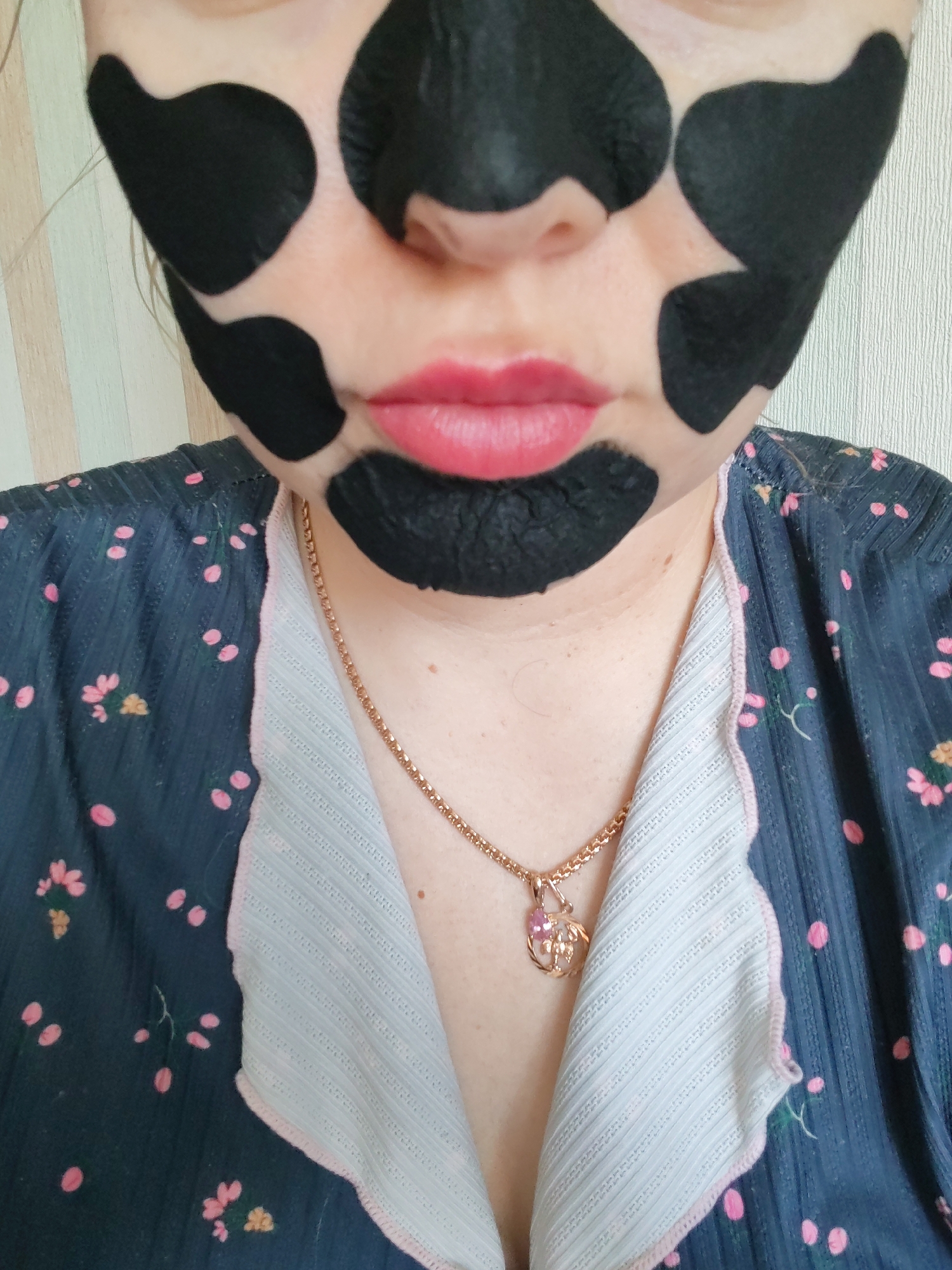 The image displays a close-up view of a person, likely an adult female, partially obscured by a facial mask, in an indoor setting, possibly in Izhevsk, Russia. Her face is covered with irregular, dark, matte black patches, giving her complexion a spotted appearance, reminiscent of a cow print or a playful application of a peel-off mask. Her lips are painted a vibrant pink, contrasting with the dark mask.
She is wearing a dark blue garment, possibly a robe or shirt, patterned with small pink and yellow flowers. Beneath this, a light-colored, ribbed fabric is visible, forming what appears to be a collar or an undershirt. A gold-toned chain necklace with a small, ornate pendant hangs around her neck. The pendant features a stylized animal, possibly a cat or fox, adorned with a small pink gemstone.
The background consists of a light-colored wall with faint vertical patterns, suggesting a domestic environment. The activity appears to be a personal care routine, specifically applying a facial mask. The expression is neutral, as much of the face is covered by the mask. No text is visible in the image.