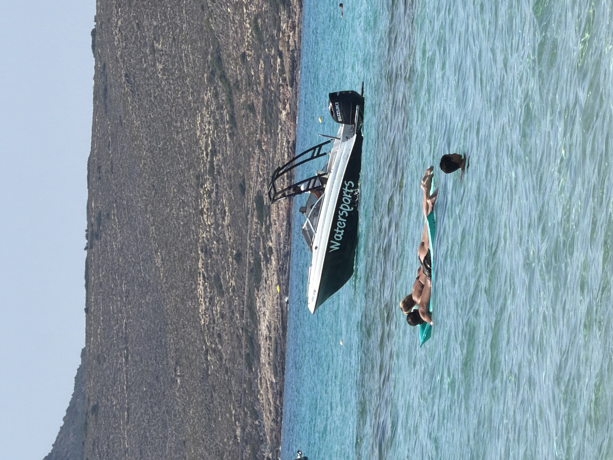 A man and a woman are lying on a green inflatable raft in clear turquoise water. The man is on his side, facing the woman, who is lying on her stomach with her head resting on her arms. Both are wearing swimwear. To the left of the raft, a young boy is swimming in the water. In the background, a white motorboat with "Watersports" written on its side is visible. The boat has a "SUZUKI" outboard motor. Beyond the water, a rocky, arid hillside with sparse vegetation is visible under a clear blue sky. The scene appears to be a coastal area, likely in Greece, given the characteristic landscape.
