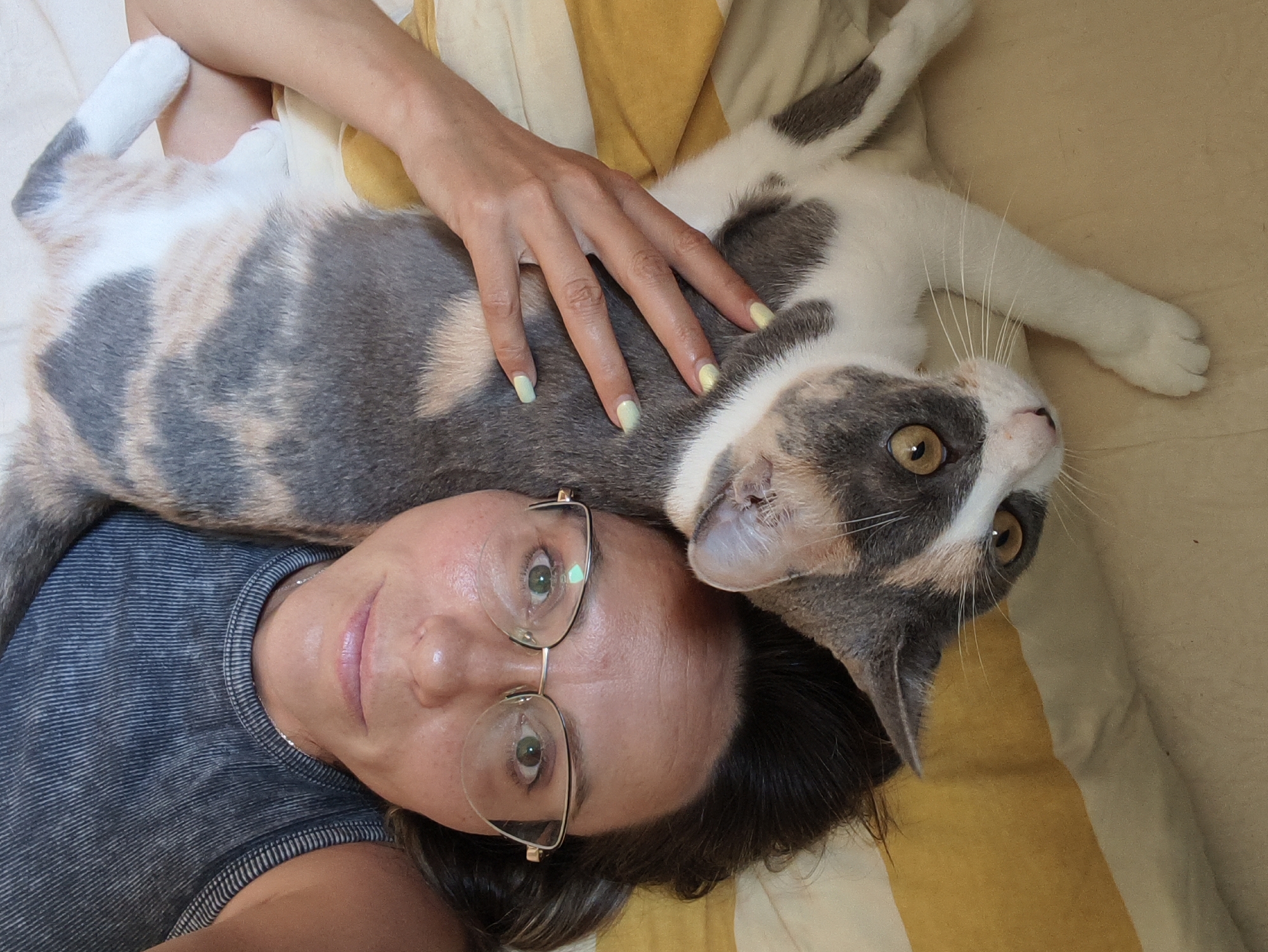 This is a close-up, overhead, portrait-style selfie taken by a person lying down, with a calico cat resting on their chest and head. The person is wearing a dark, striped tank top and eyeglasses. Their eyes are green and wide open, and they are smiling slightly. The cat is predominantly white with patches of grey, tan, and black. Its left paw is extended and resting on the person's upper chest, and its right paw is tucked underneath its body. The cat's head is turned towards the camera, with large, amber eyes. The background consists of a textured surface, possibly a bed or couch, with horizontal stripes of yellow and off-white. The image is well-lit, with soft shadows. The image does not contain information about the city of Sochi, Russia.