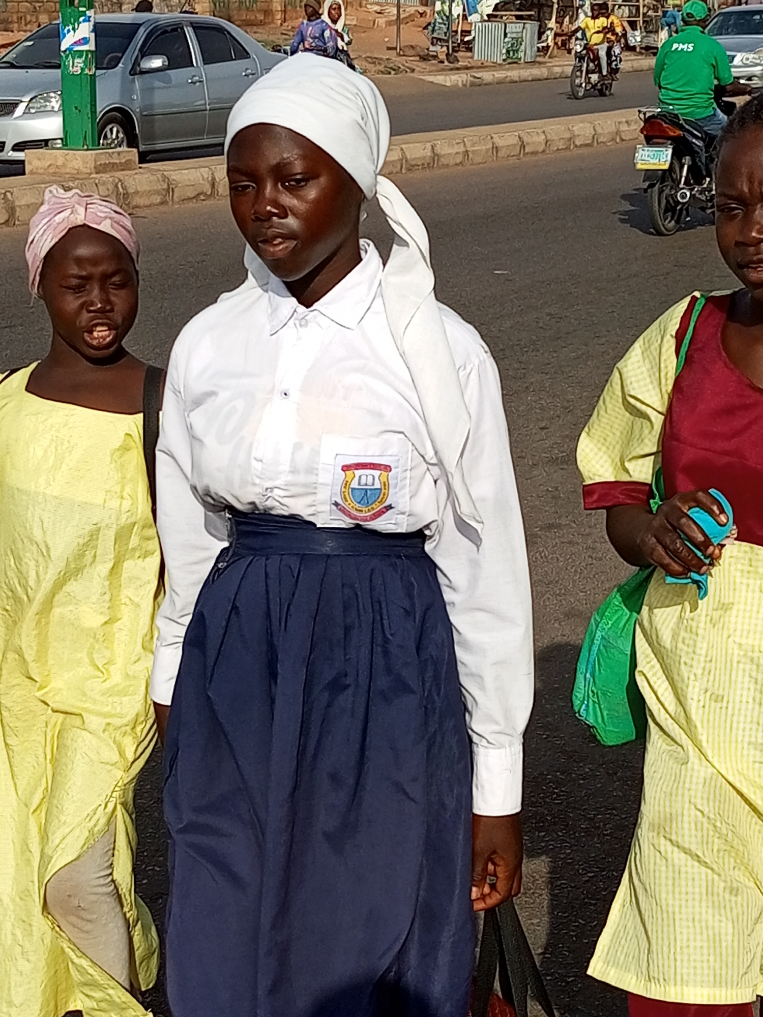 Three young females are walking on a paved road in the city of Gombe, Nigeria. The female in the center is wearing a white long-sleeved shirt, a navy blue pleated skirt, and a white headscarf. She has a school emblem on her shirt pocket. To her left, a female in a yellow tunic and a pink headscarf is walking with her mouth open. To the right of the center female, a third female, partially visible, is wearing a yellow and white checkered garment with a maroon overlay and carrying a green bag. In the background, there is a silver car, a motorcycle with two passengers, and various buildings. The road appears to be a busy urban street.