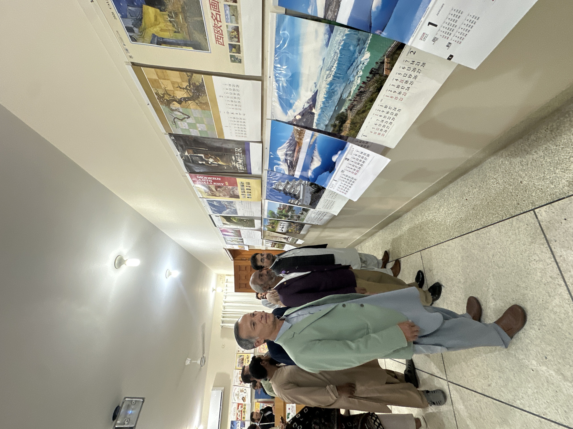 The image depicts a group of men standing in a hallway, presumably in Quetta City Tehsil, Pakistan. They are lined up along a wall covered with calendars and posters. The men are dressed in a variety of attire, including suits and traditional Pakistani clothing. They appear to be looking at the wall displays. The hallway is brightly lit with overhead lighting. The floor is tiled.