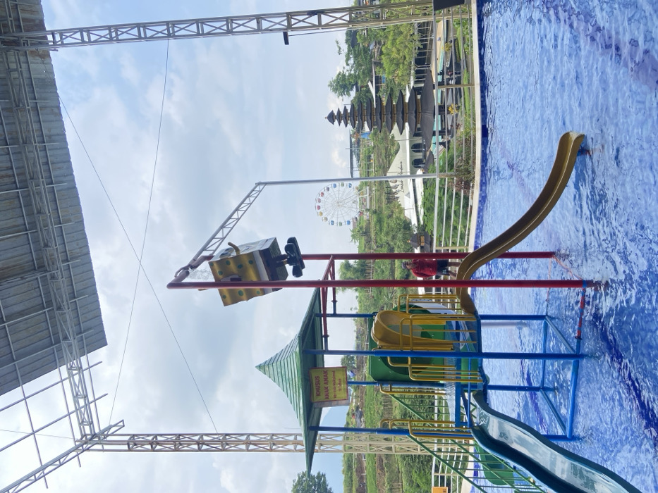 A water park features a pool with a blue slide and an overhead structure that resembles a die with a metal arm extending to a figure on top. A Ferris wheel and a multi-tiered building are visible in the background, suggesting the park's various attractions. The pool's clear, blue water and the slides encourage a playful atmosphere. The scene suggests an outdoor recreational space for families.