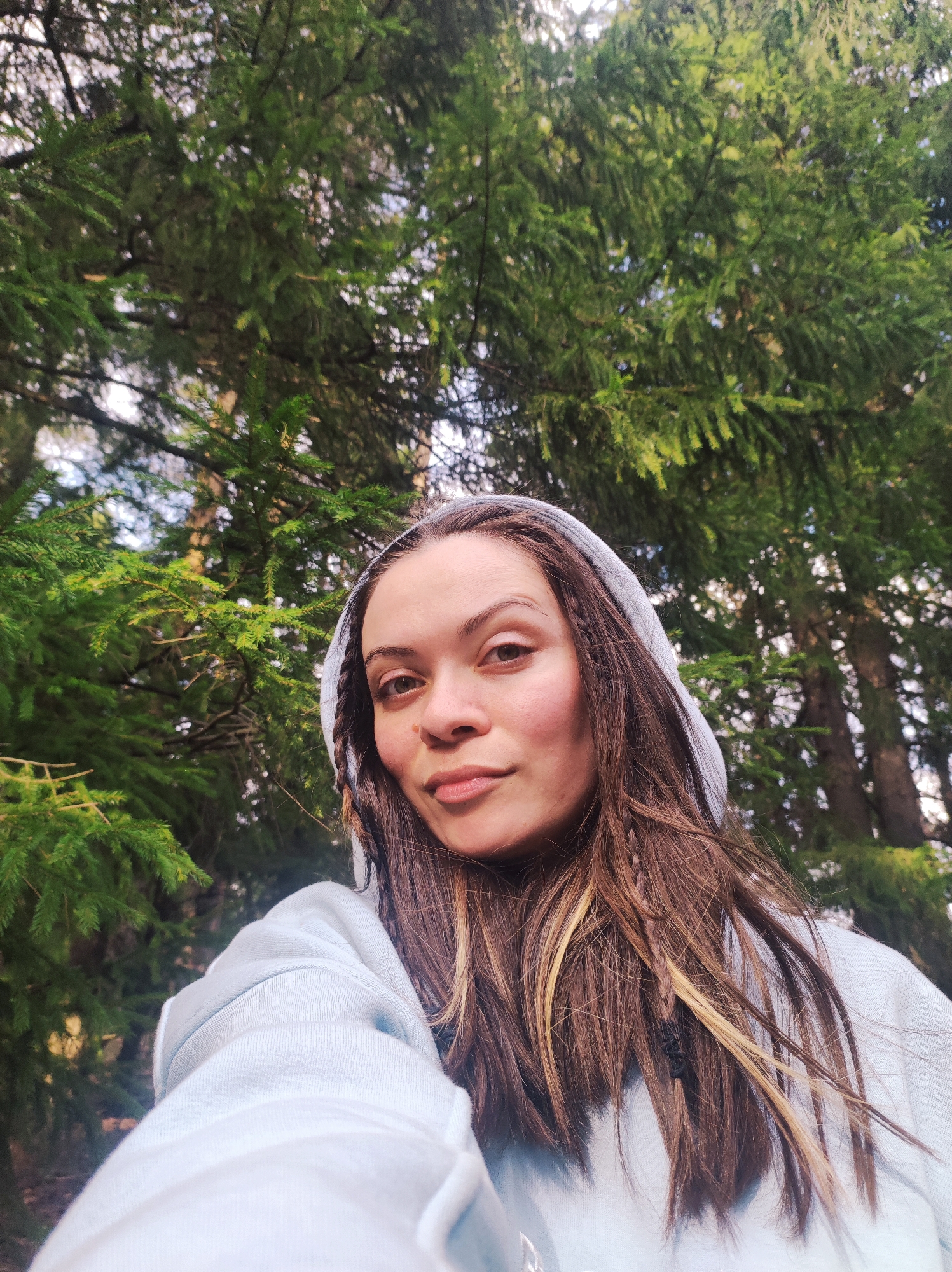 A young woman takes a selfie in a forest setting in Фоки, Russia. She is positioned in the foreground, looking directly at the camera with a calm and natural expression. Her long, dark brown hair, featuring noticeable blonde or caramel highlights and some small dark braids, frames her face and falls over her shoulders. She wears a light blue or grey hooded sweatshirt, with the hood pulled up over her head. The fabric of the sweatshirt appears soft and textured.
The background is dominated by dense evergreen trees, likely spruce or fir, with their branches and needles filling the upper two-thirds of the frame. The perspective is from below, looking up into the canopy, where patches of a bright, possibly overcast sky can be seen through the foliage. The lighting suggests it is daytime, with diffused light filtering through the trees, casting soft illumination on the scene. There is no visible text in the image. The overall impression is one of tranquility and connection with nature.