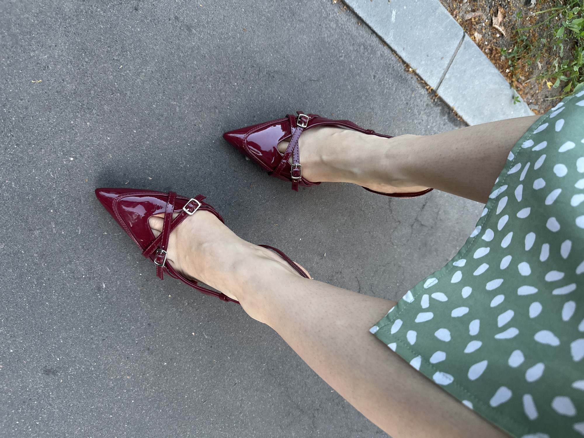 This is a top-down, medium shot image of a person's legs and feet, standing on asphalt. The person is wearing a green dress with a white polka dot pattern. They are also wearing pointed, patent leather, burgundy high-heeled shoes with crisscross straps and buckles. The shoes appear to be slingbacks, as the heel is open. The setting is outdoors, with a sidewalk made of grey concrete slabs to the right of the asphalt. Some sparse vegetation and dried leaves are visible along the edge of the sidewalk. The lighting suggests it is daytime, possibly sunny, given the reflections on the patent leather shoes. There is no visible text in the image. The overall mood is casual and summery.