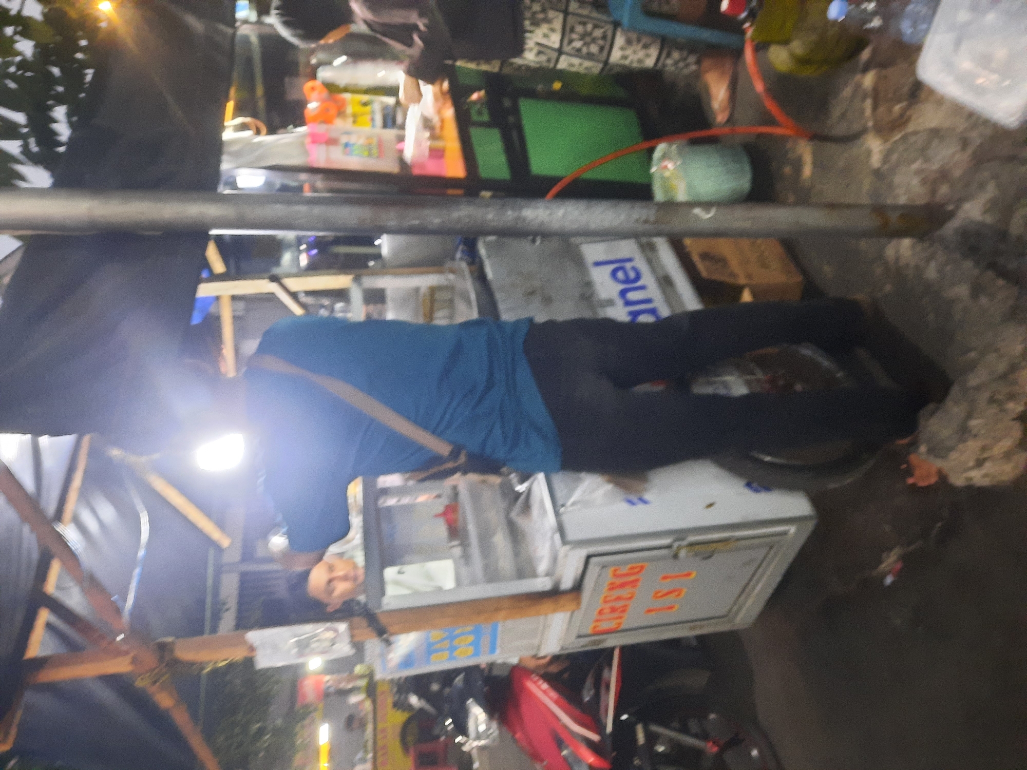 An outdoor night scene depicts a food cart with a person in a blue shirt facing away from the camera, appearing to be attending to customers. The food cart has a white exterior with orange lettering that reads "CIRENG ISI". Another person, wearing a dark top and patterned pants, is partially visible to the right, suggesting a transaction or interaction at the cart. The background shows other food stalls or vendors, a red motorcycle, and foliage, indicating a busy street food environment. The lighting is dim, with artificial lights from the stalls illuminating the scene. The overall impression is of a lively, informal market setting, likely in a bustling urban area of North Jakarta, Indonesia.